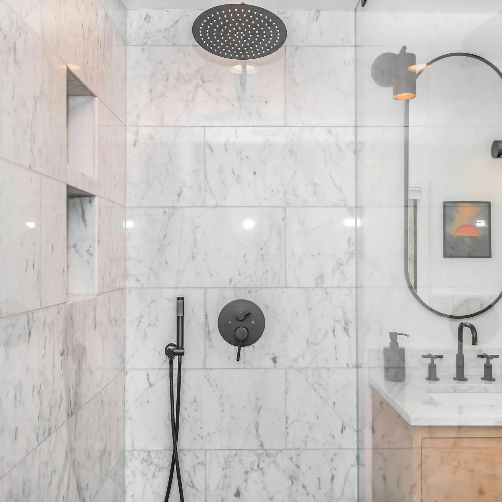 Modern bathroom shower with marble tiles, rainfall showerhead, control knobs, and a handheld showerhead. A glass panel separates the shower from the vanity area with a round mirror and black faucet.