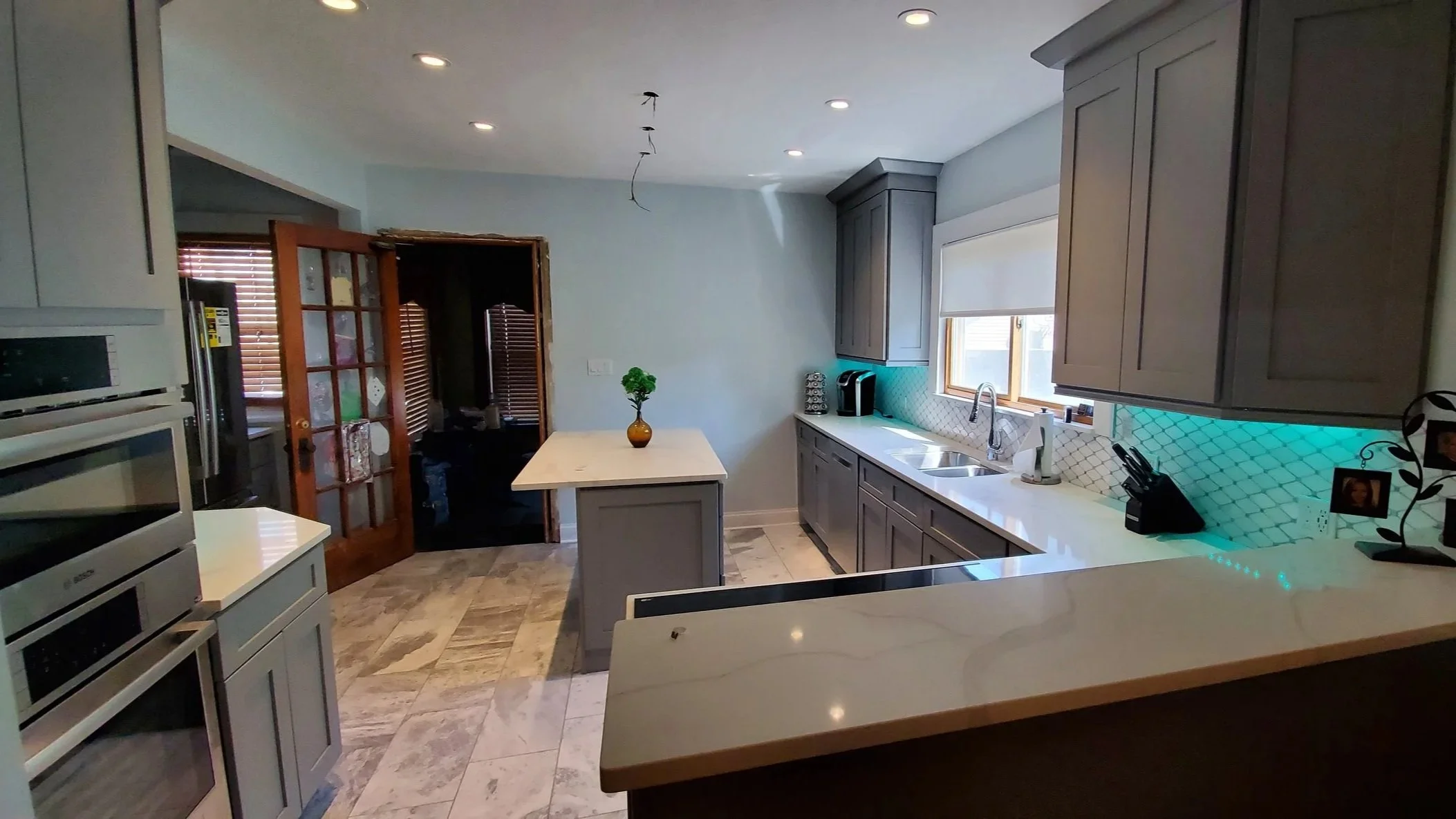 Kitchen with gray cabinets, marble floor, and a white countertop. There is a small gray island with a vase and a green plant, a double sink under a window, and some kitchen appliances. A sliding glass door with wooden frames leads to another room. Recessed ceiling lights and under-cabinet lighting are present.