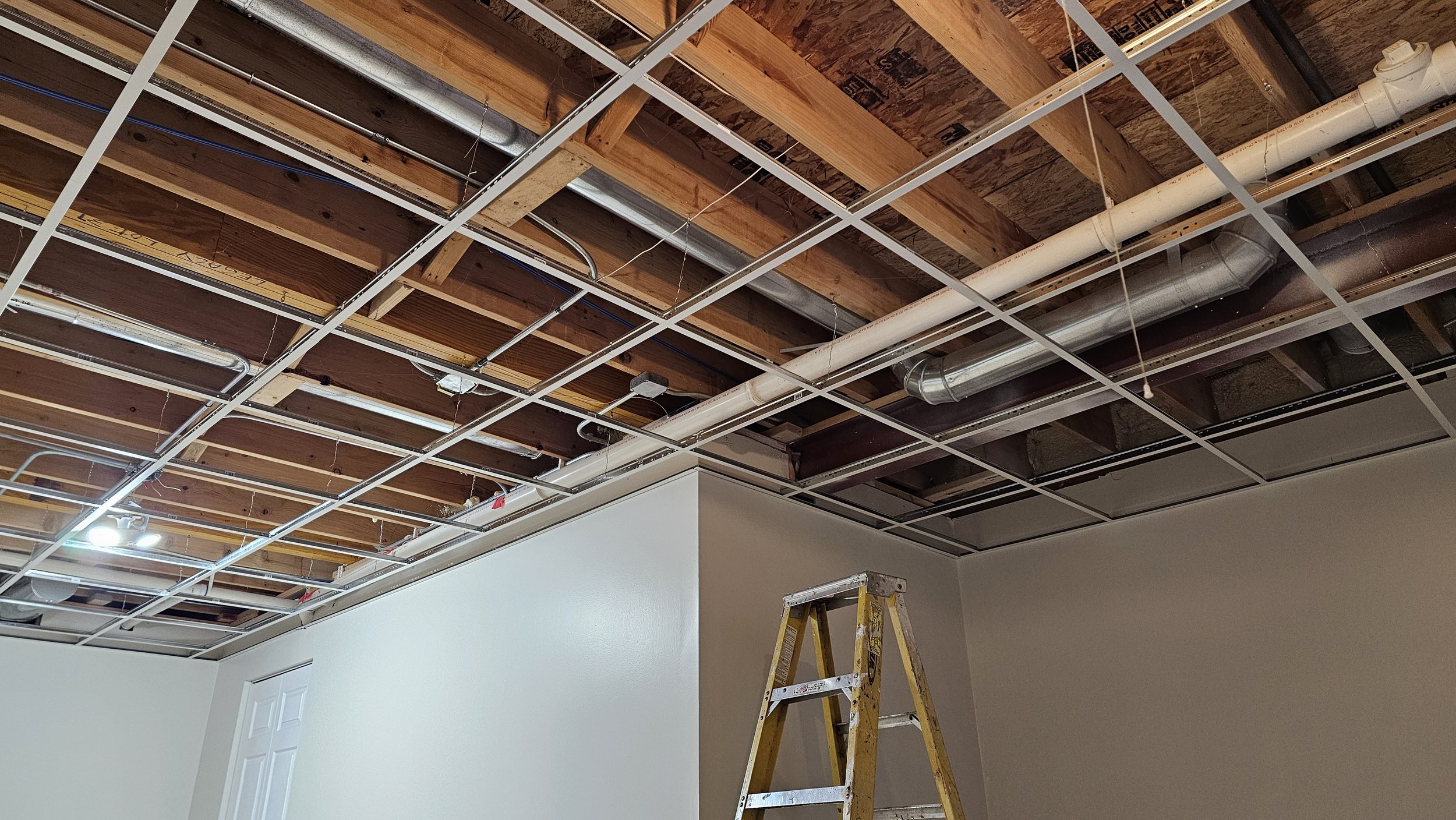 Room under renovation with exposed ceiling framework, metal ducts, electrical wiring, and a yellow ladder.