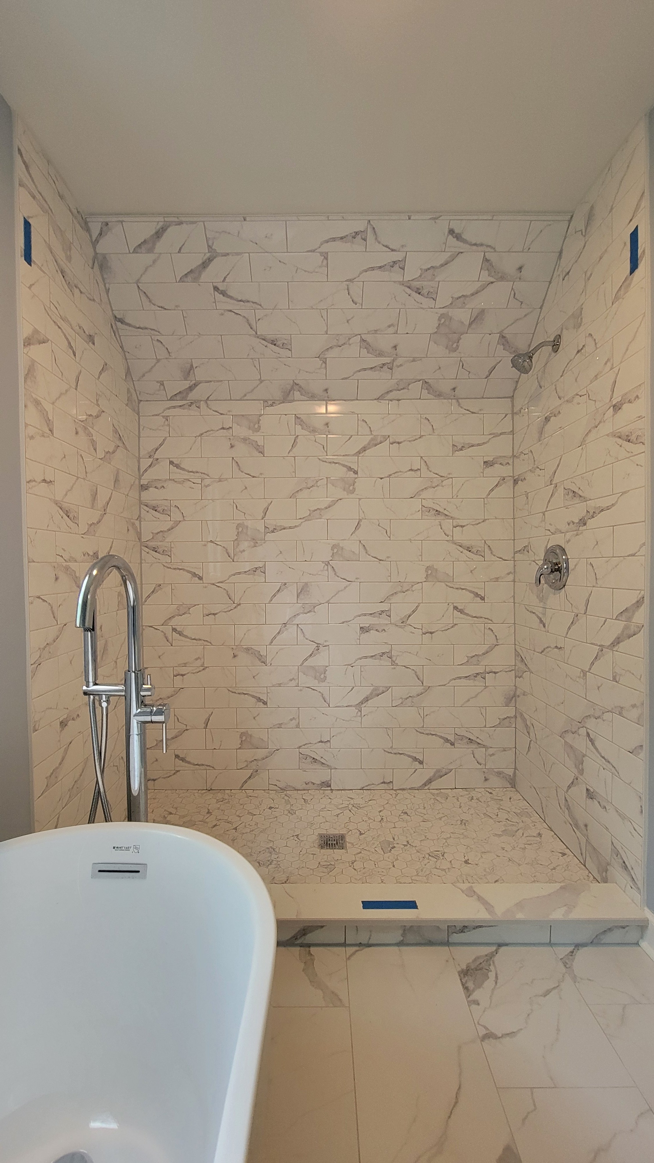 Empty shower with beige and gray marble tile walls, a small hexagonal tile floor, a ceiling shower head, and a wall-mounted control valve. A bathtub with a chrome faucet is visible in the foreground.