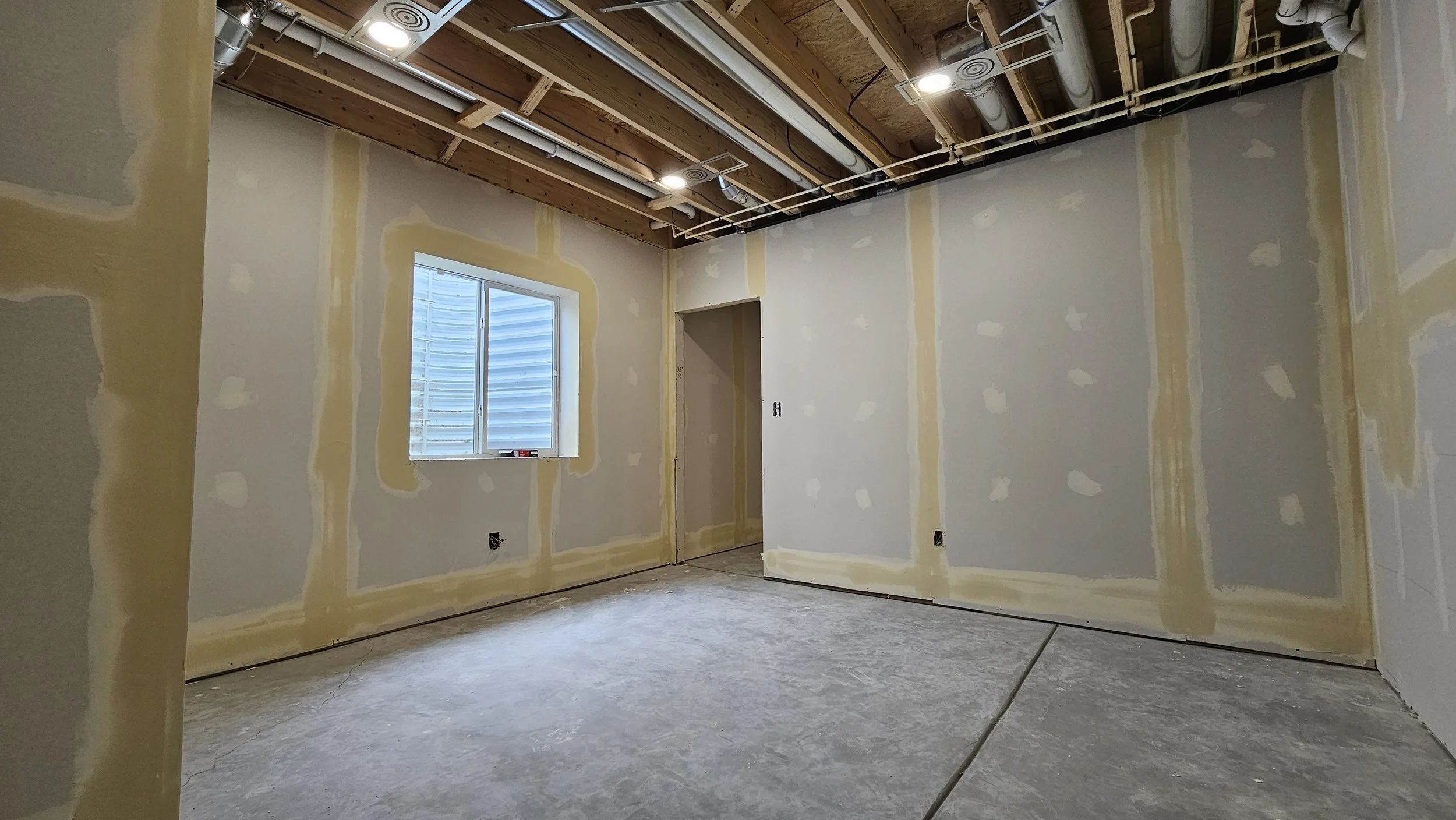 An unfinished room with drywall installed, a window, and exposed ceiling framing with electrical and ductwork.