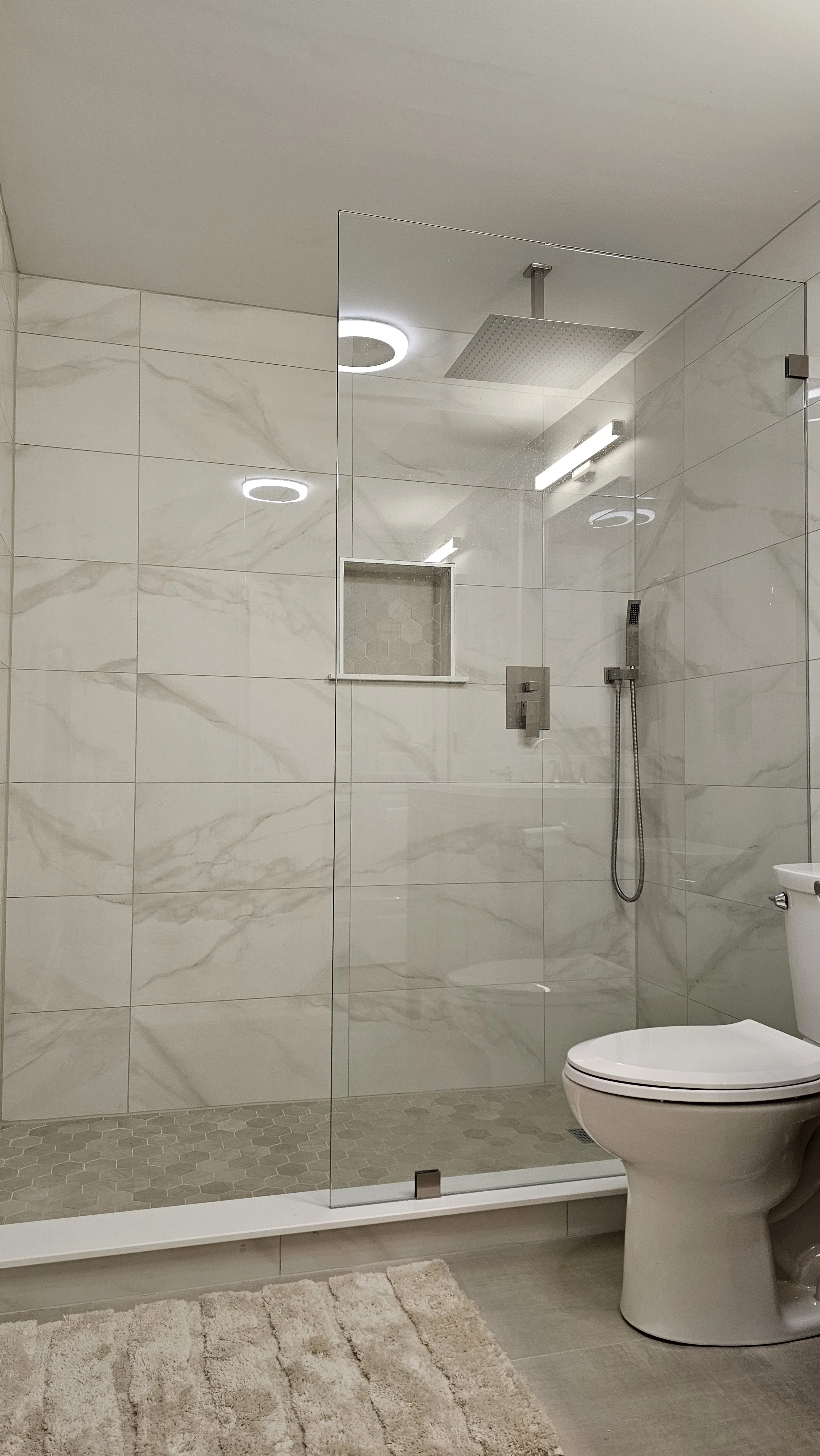 Modern bathroom with a large glass-enclosed shower, marble tiles, a rain shower head, and a white toilet. A beige bath rug is on the floor outside the shower.