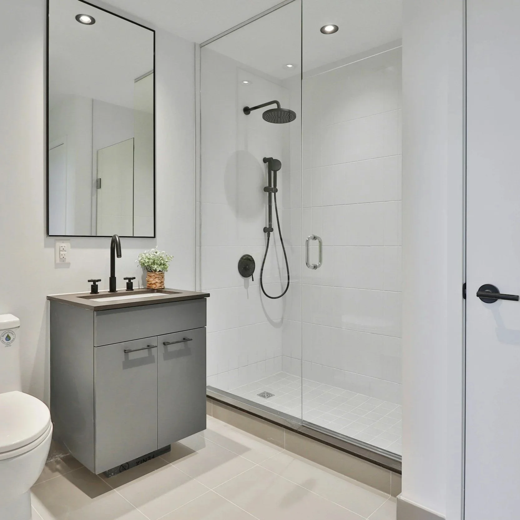 Modern bathroom with walk-in shower, gray vanity with sink, mirror, and a small potted plant, with white tiled walls and floor.