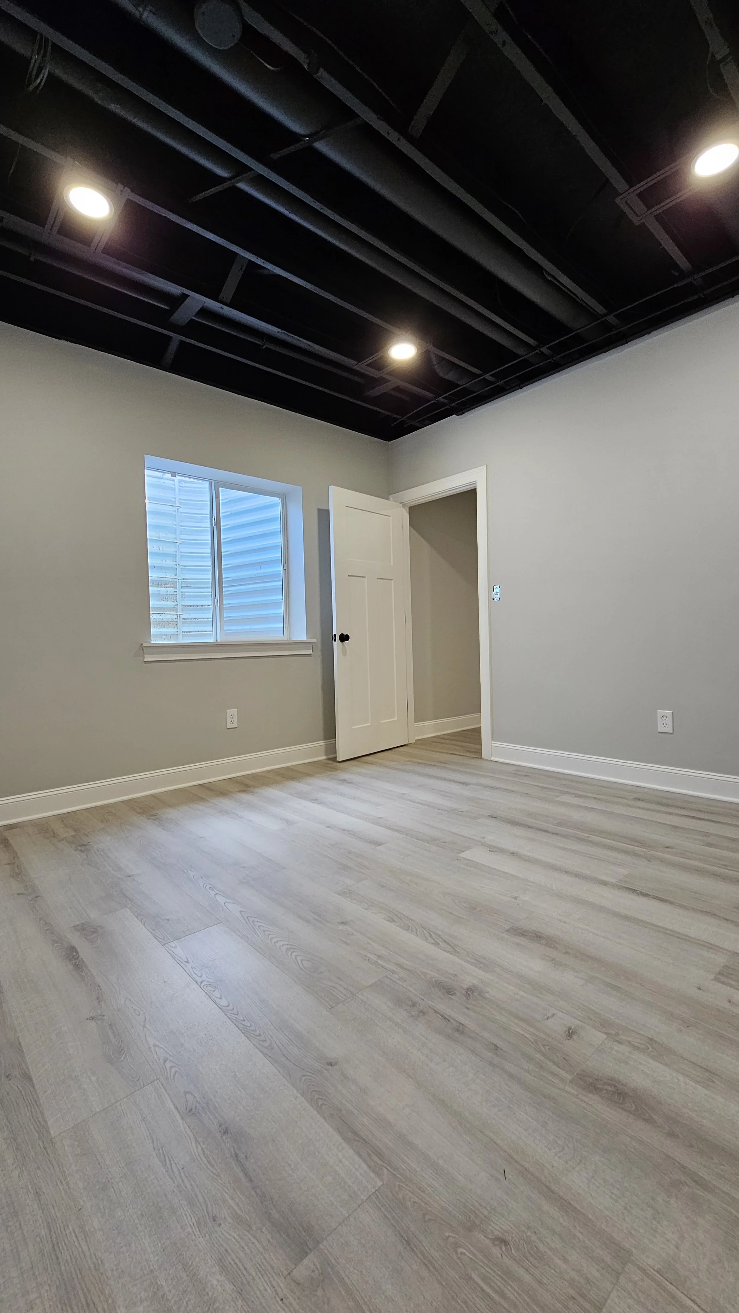 Empty room with light-colored wood flooring, light gray walls, a small window, and a white door, with a black ceiling that has recessed lighting.