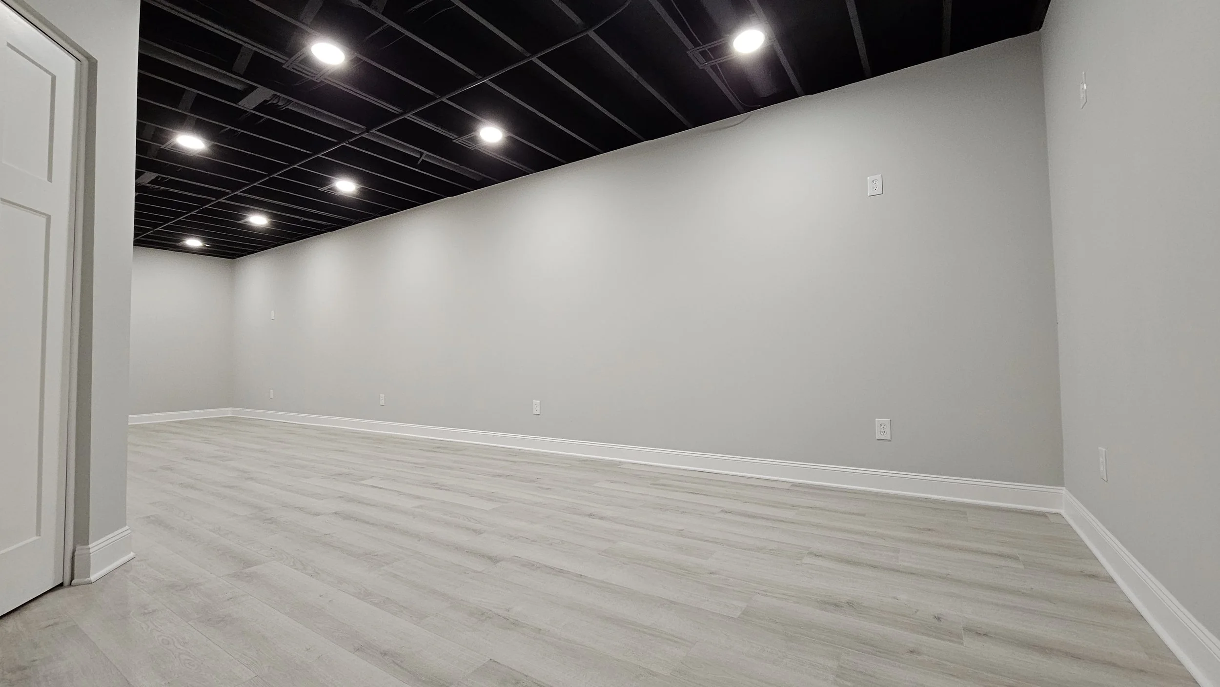 Empty room with white walls, light wood flooring, and black ceiling panels with built-in lights.