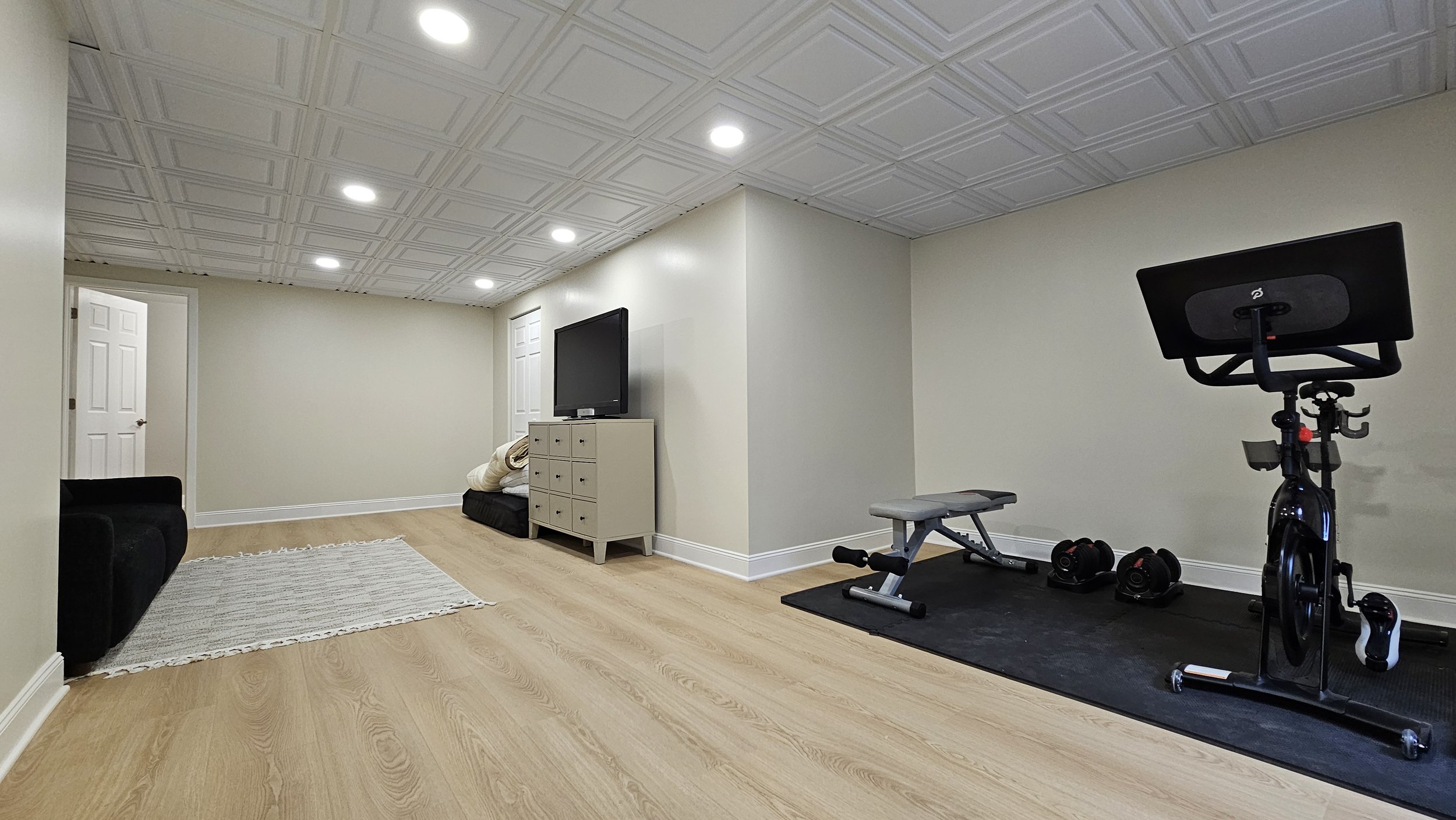 A basement gym with workout equipment, a flat-screen TV on a white dresser, a black armchair, a small rug, and a beige wall with white doors. Exercise bike, dumbbells, and a weight bench are on a black mat.