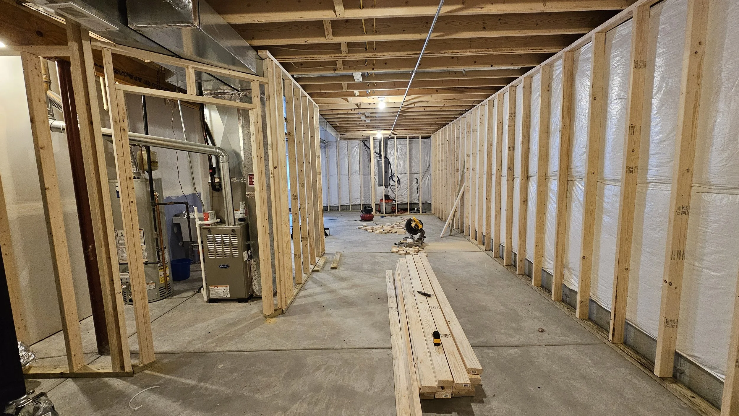 Interior view of a construction site with exposed wooden framing, insulation on the right wall, and construction tools on the floor, including a circular saw and a measuring tape.