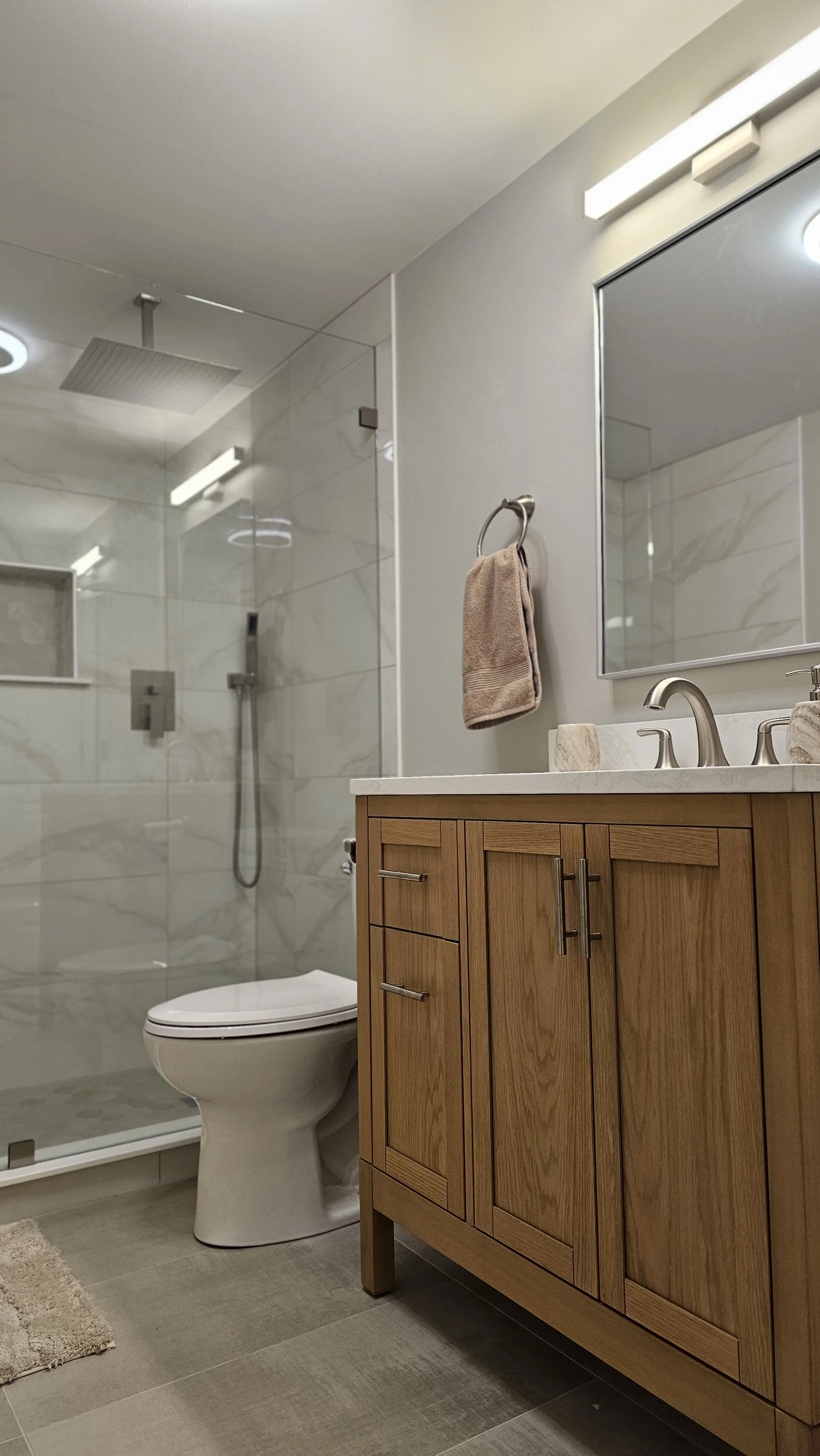 Modern bathroom with a glass shower enclosure, white toilet, wooden vanity with a marble countertop, and a large mirror above the sink. There is a beige towel hanging on a ring on the wall.