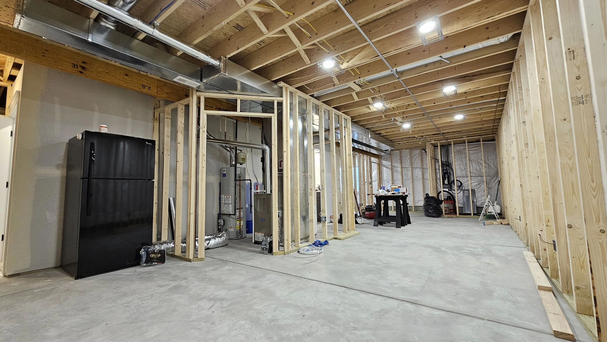 Interior of a building under construction with exposed wooden framing on walls and ceiling, concrete floor, and ceiling lights installed. Construction tools and materials are scattered around, including a black refrigerator, water heater, and insulat