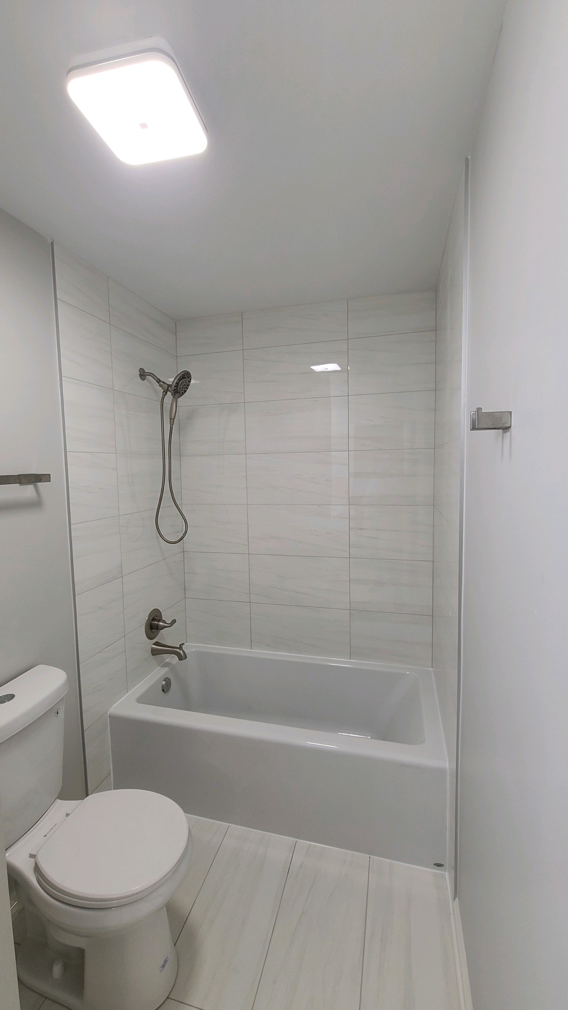 Modern bathroom with a white toilet, bathtub, and showerhead, white tiled walls, and ceiling light.