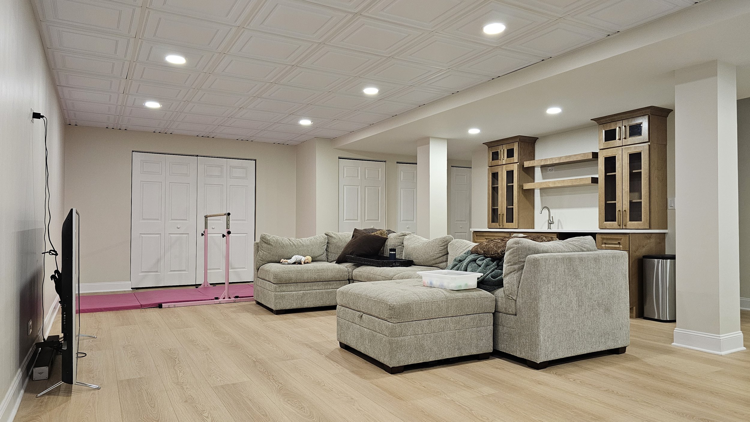 A living room with a beige sectional sofa, a TV on a stand, a pink exercise treadmill, a wooden built-in cabinet, a small sink, and a trash can. The ceiling has recessed lighting and the floor is light wood.