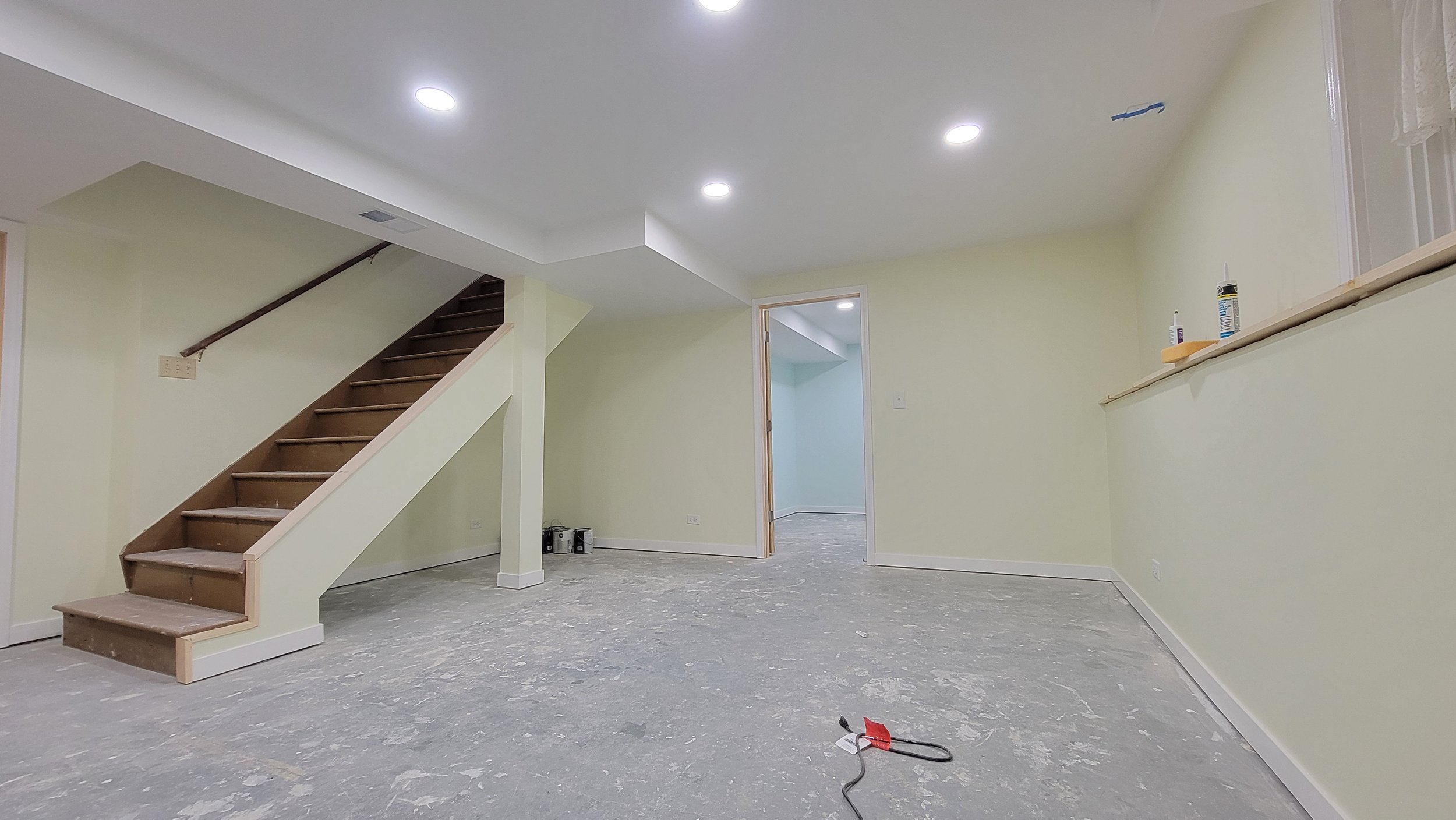 Empty room with yellow walls and ceiling lights, unfinished floor, staircase on the left, and a door opening to another room in the background.