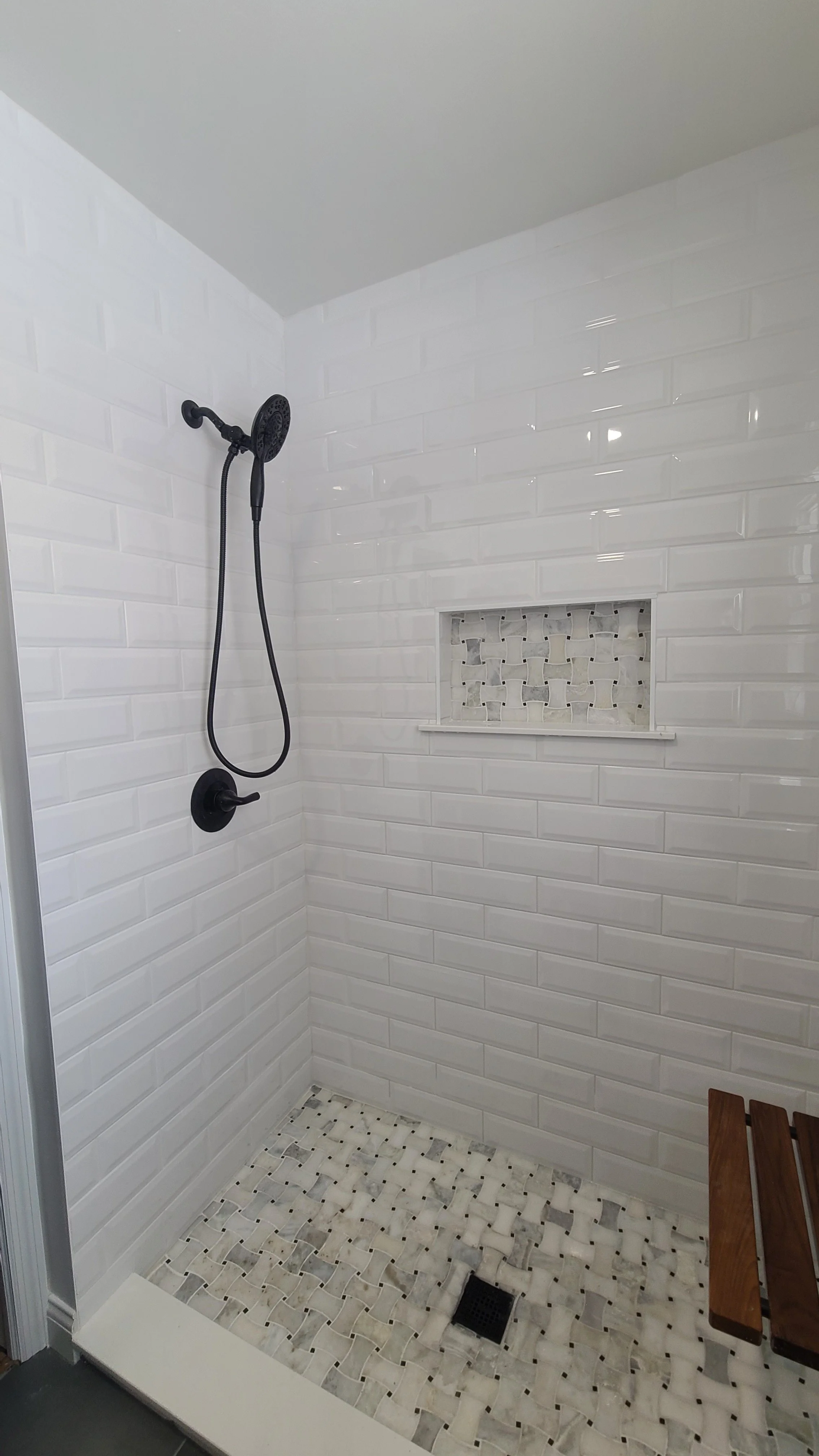 A modern walk-in shower with white subway tile walls, a built-in niche with a decorative mosaic tile insert, a black showerhead and handle, a black floor drain, and a wooden bench.