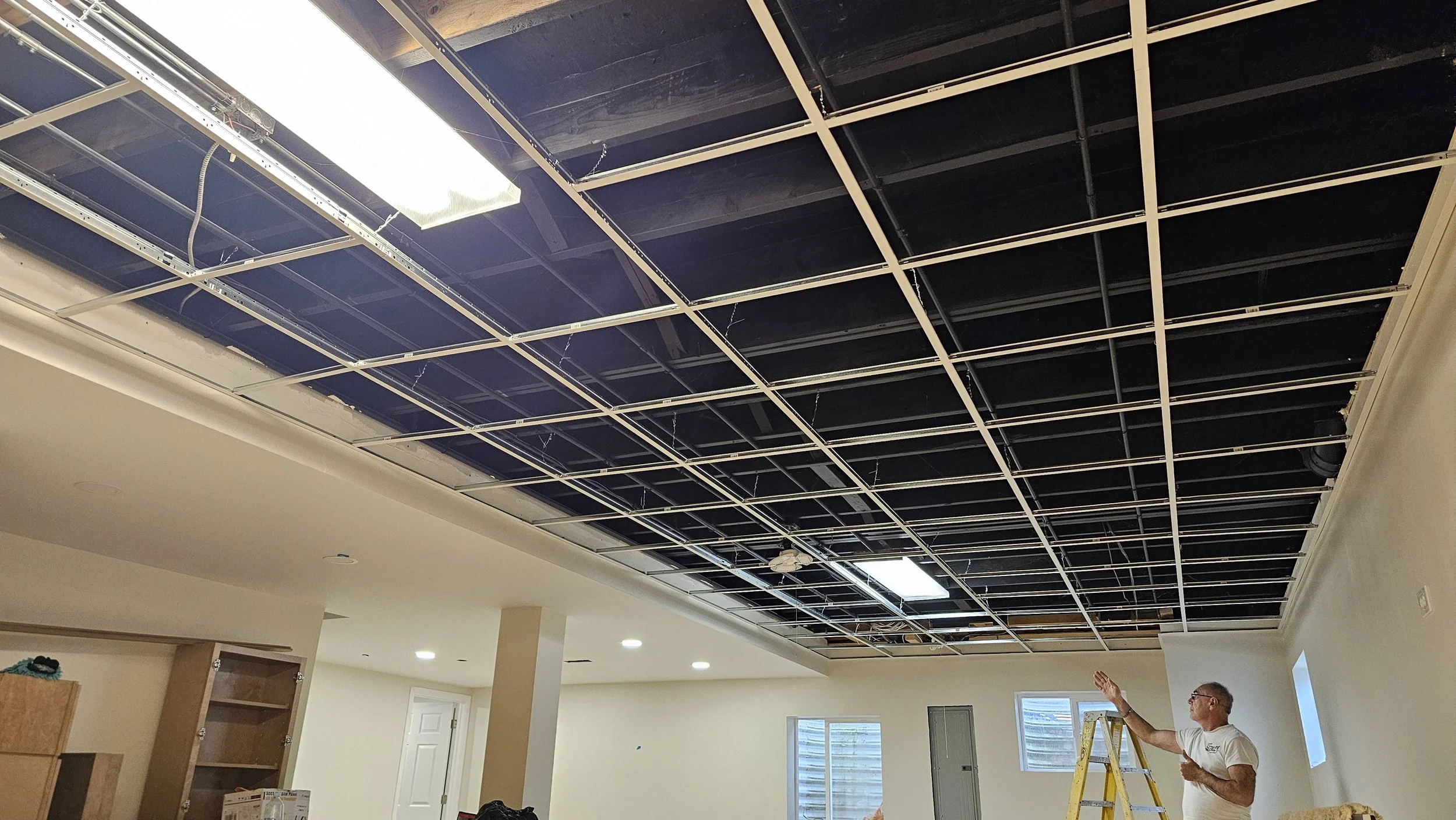 A man standing on a yellow ladder pointing upward at a ceiling under renovation, with exposed black electrical wiring and metal framework, in a room with cream-colored walls and multiple windows.