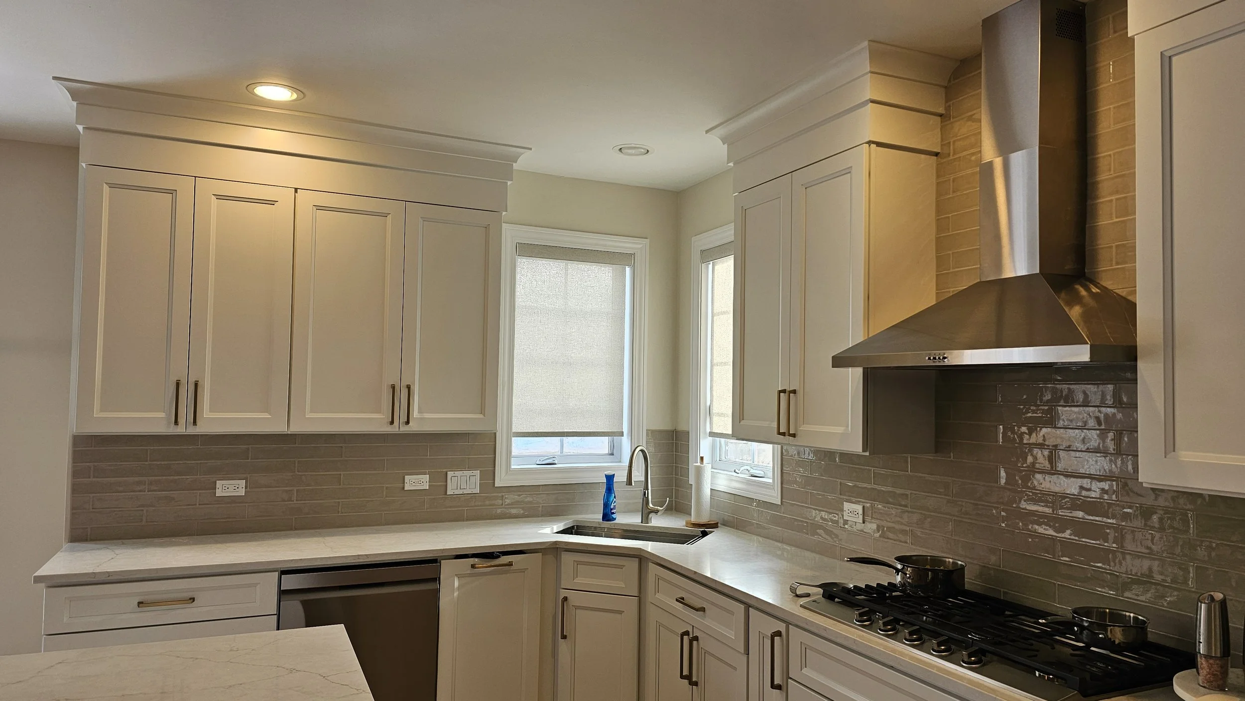 Modern kitchen with white cabinets, beige brick backsplash, stainless steel range hood, gas stove, and a window above the sink with a blue bottle.