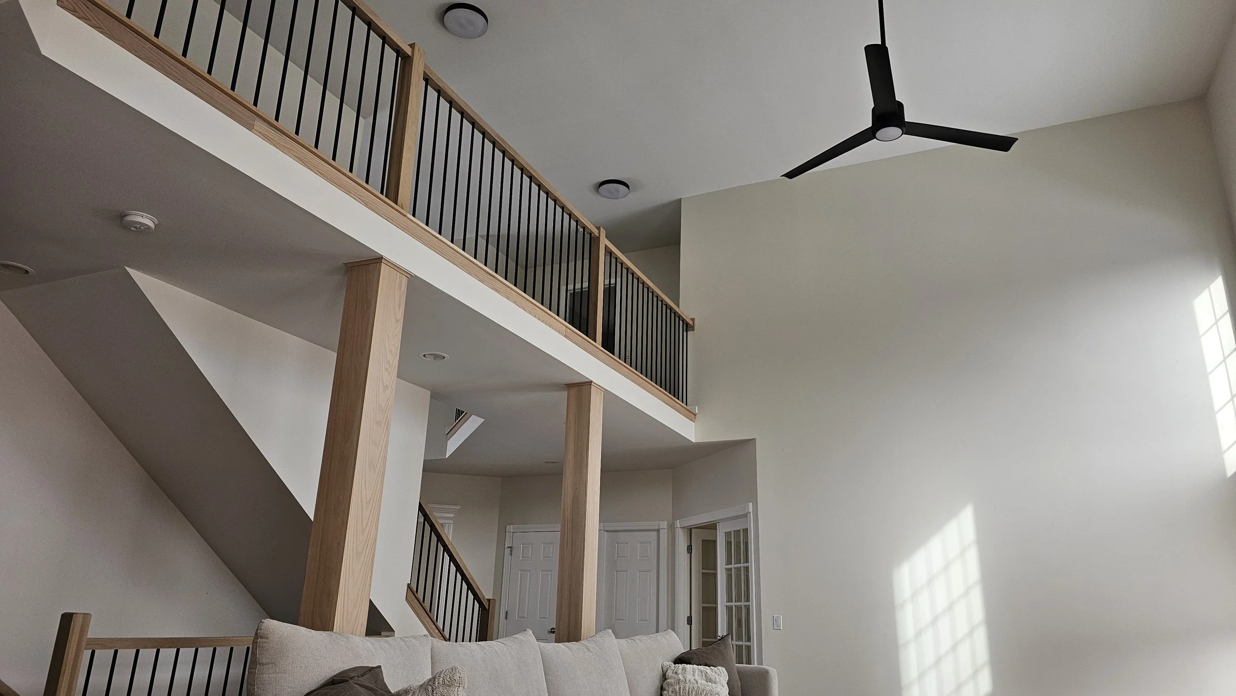 Interior of a modern two-story living room with a beige sofa, a black ceiling fan, wooden balcony railing, and large windows casting natural light on the off-white walls.