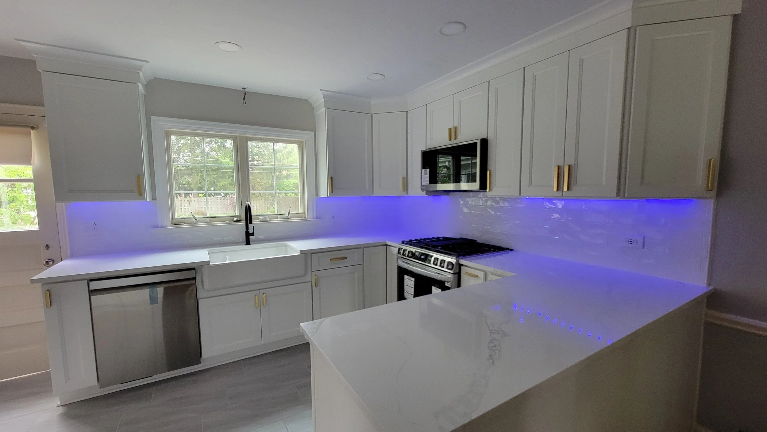 Modern kitchen with white cabinets, a large window above the sink, a stove, microwave, and blue LED accent lighting along the back splash.