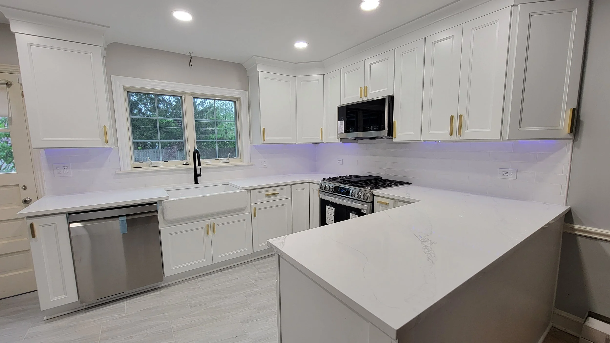 Modern white kitchen with white cabinets, a large window, black faucet, stainless steel appliances, white marble countertops, and purple LED lighting underneath cabinets.