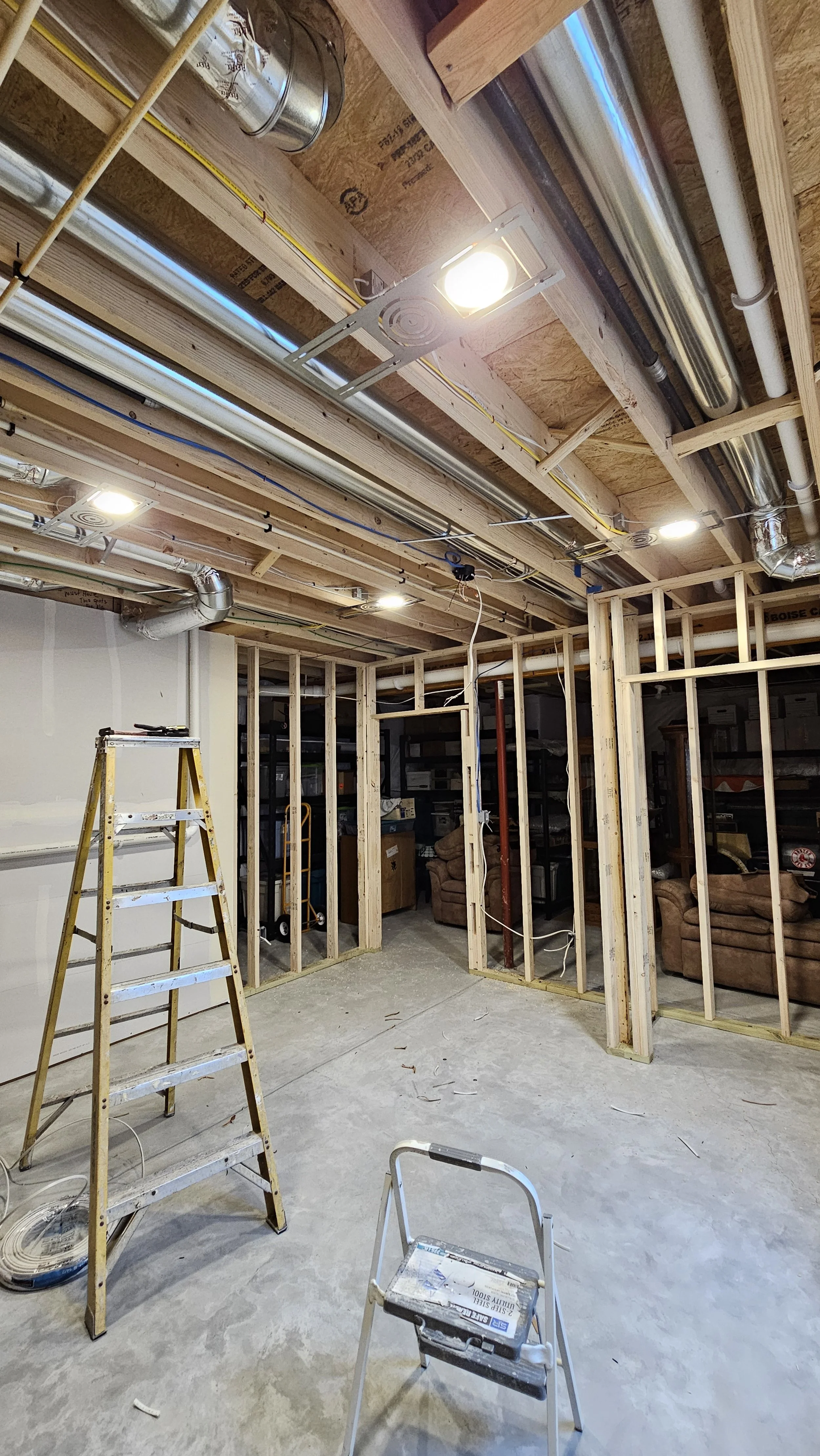 Interior of a room under construction with exposed wooden framing, ceiling with lights and ducts, and construction tools including a step ladder and small step stool on a concrete floor.
