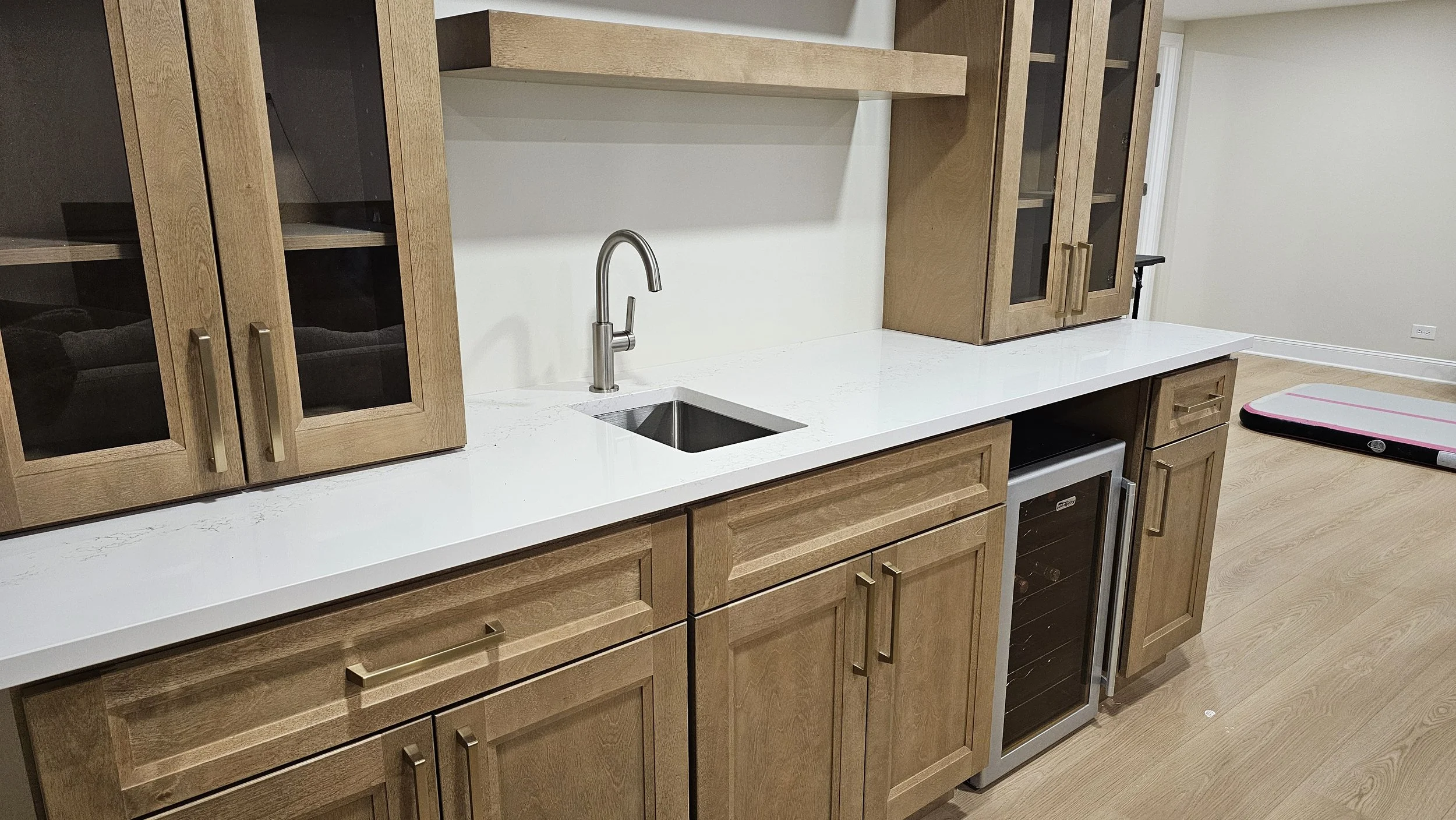 Kitchen area with wooden cabinets, a white countertop, a small sink with a silver faucet, and a mini wine cooler, with a foam roller on the floor in the background.