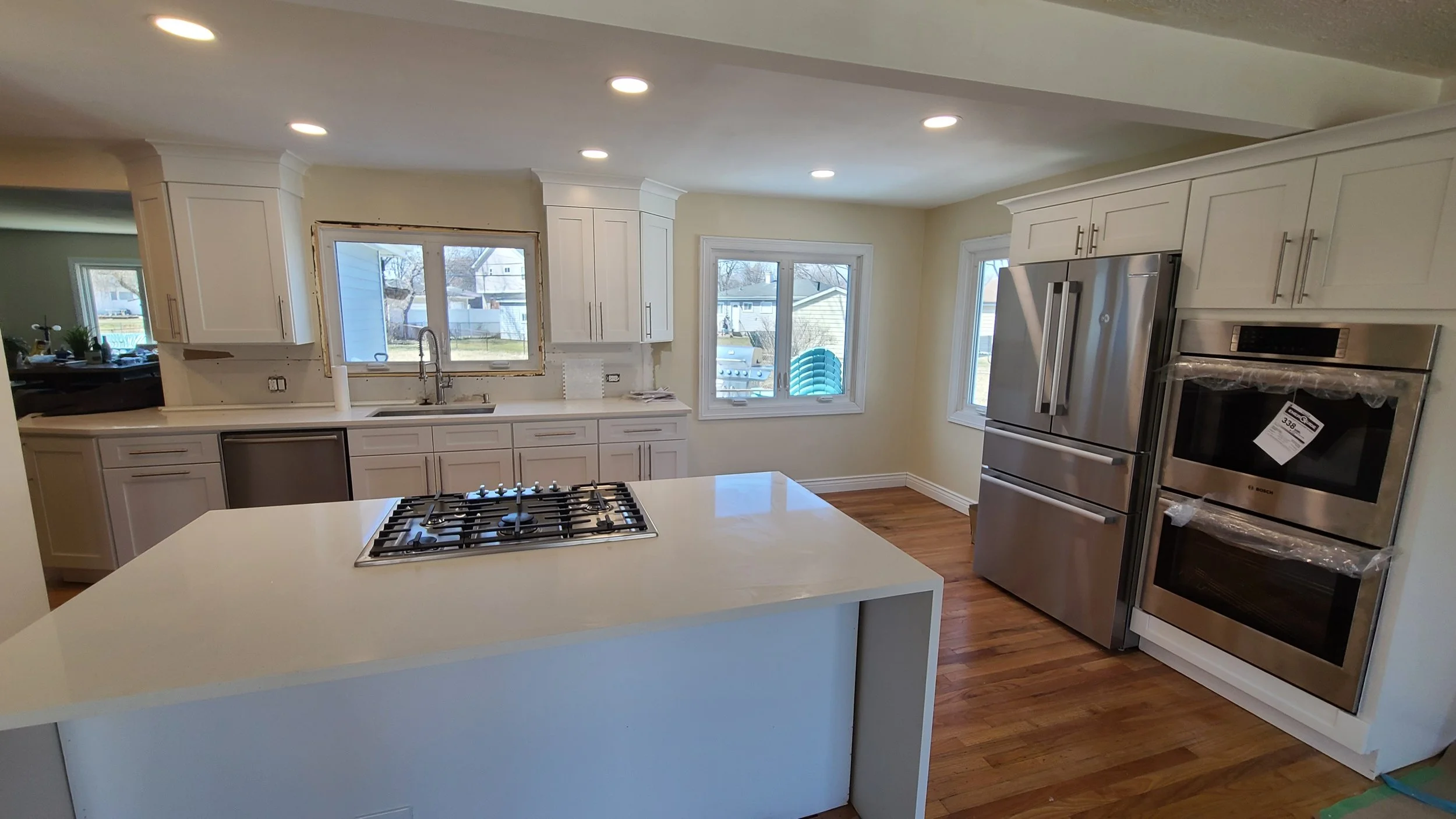 Kitchen with white cabinets, stainless steel appliances, and hardwood floors, featuring a large island with a gas cooktop and three windows providing natural light.