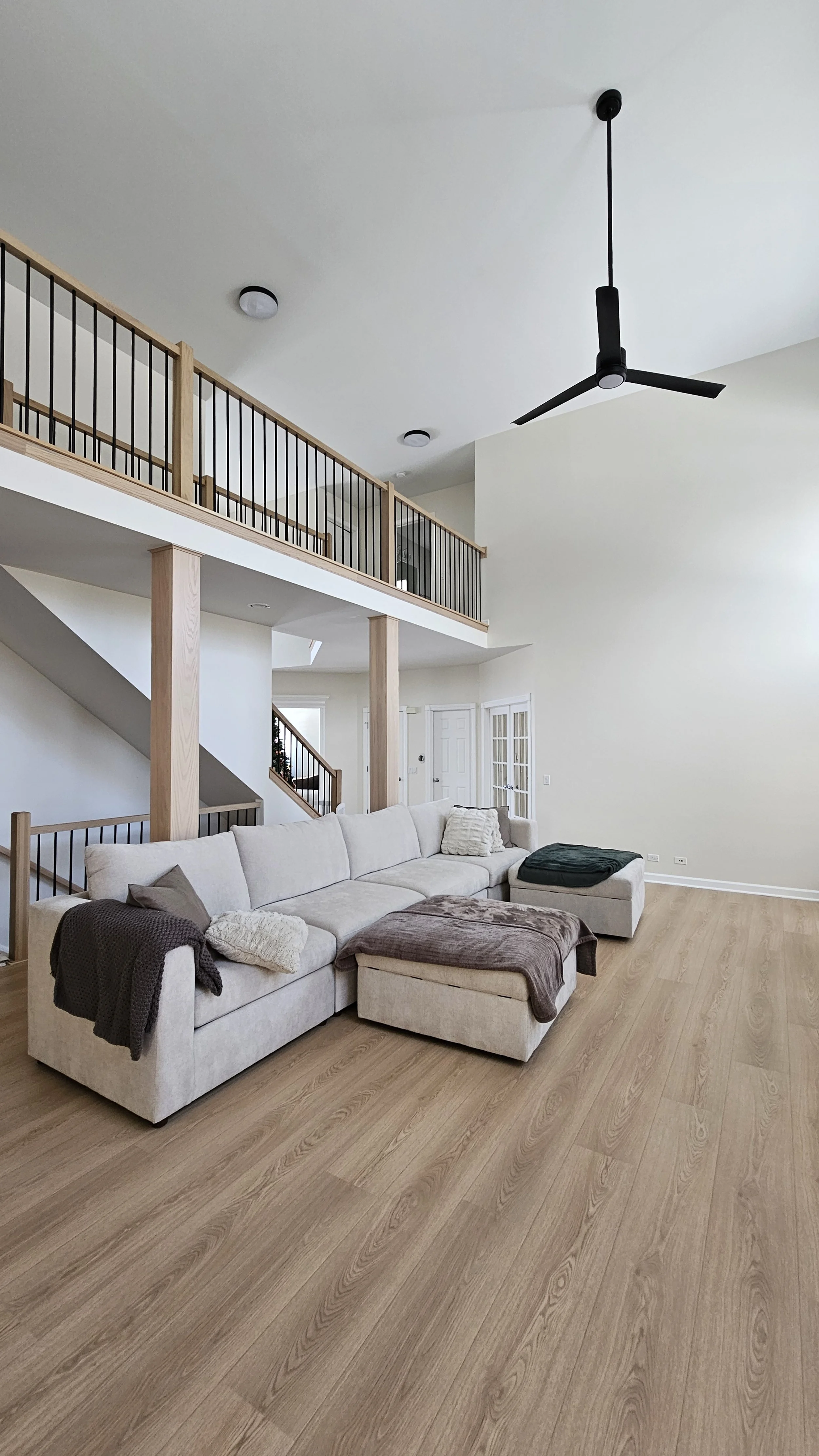 Living room with a large beige sectional sofa, wooden flooring, modern black ceiling fan, and a staircase with wooden handrails leading to a second floor.