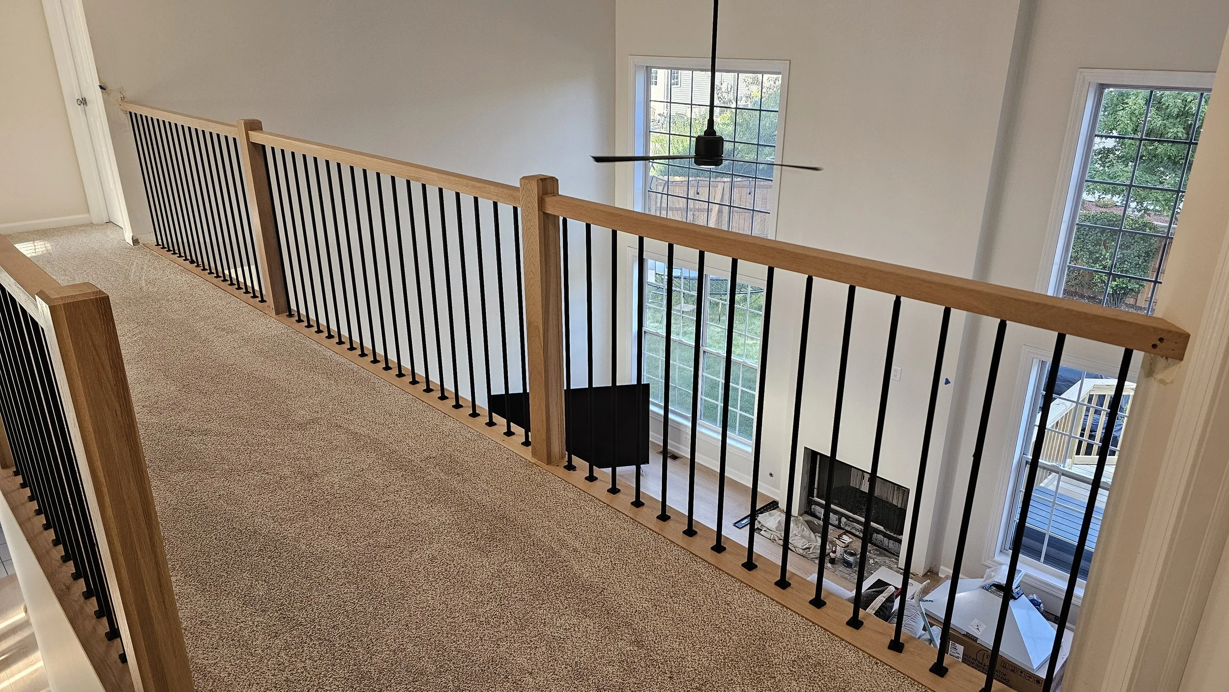 View of a second-story open hallway with a wooden and metal railing, overlooking a living room with large windows, a fireplace, and a ceiling fan.