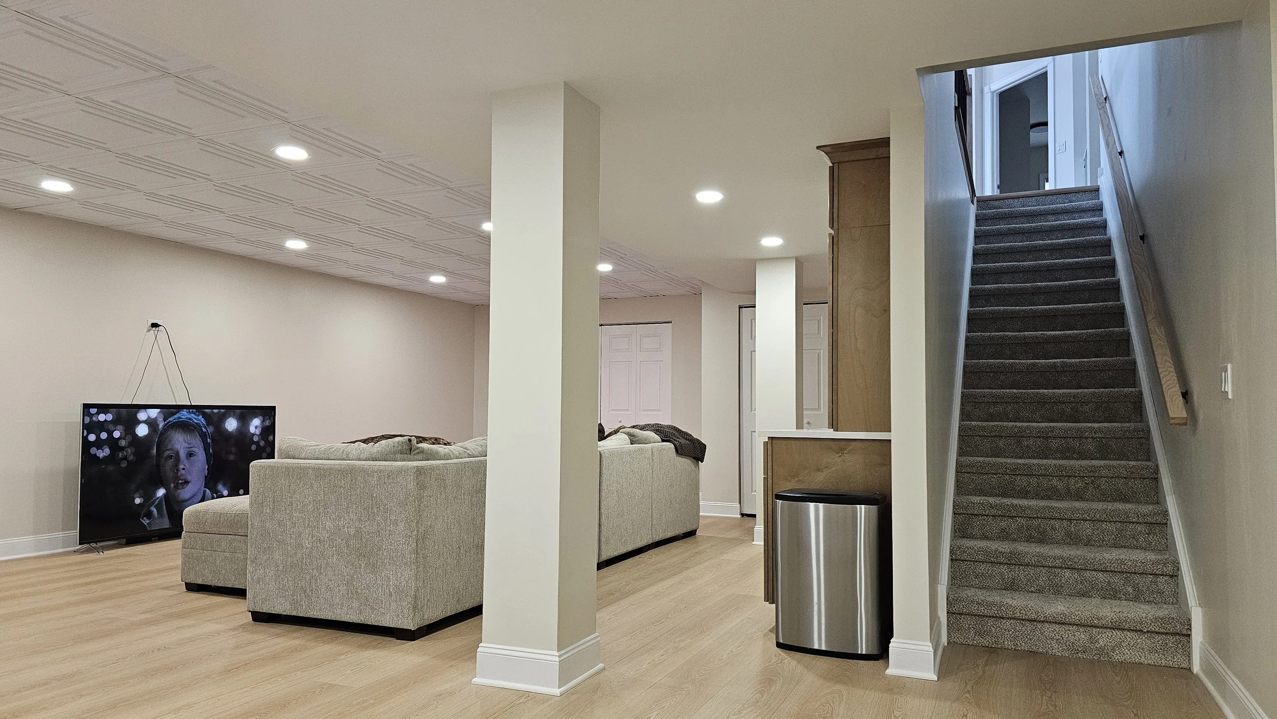 Living room with a wall-mounted TV showing a young girl, beige sectional sofas, a staircase with gray carpet leading upstairs, and light wood flooring and walls.