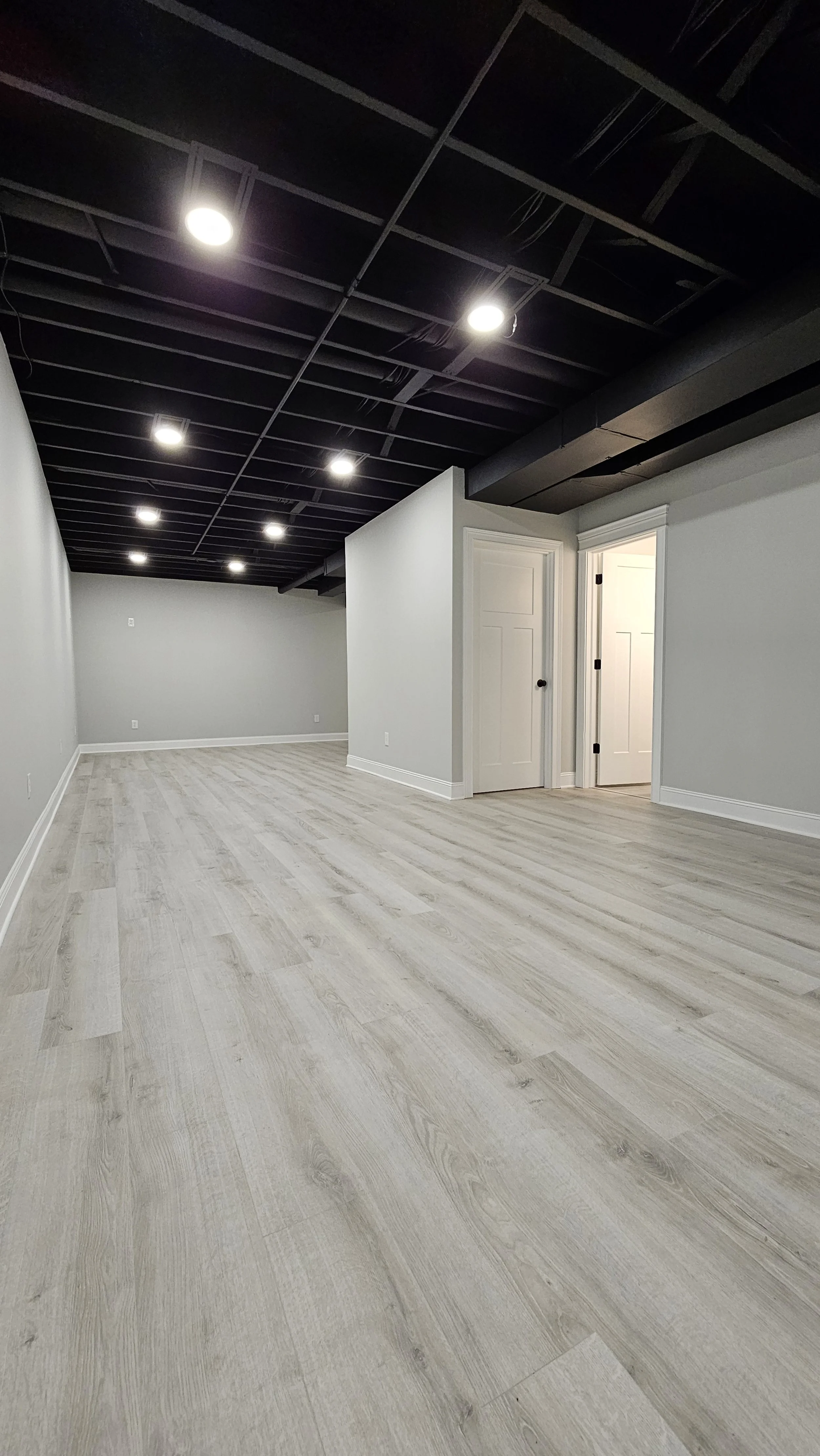 Empty basement with light wood flooring, white walls, and black ceiling with installed recessed lighting.