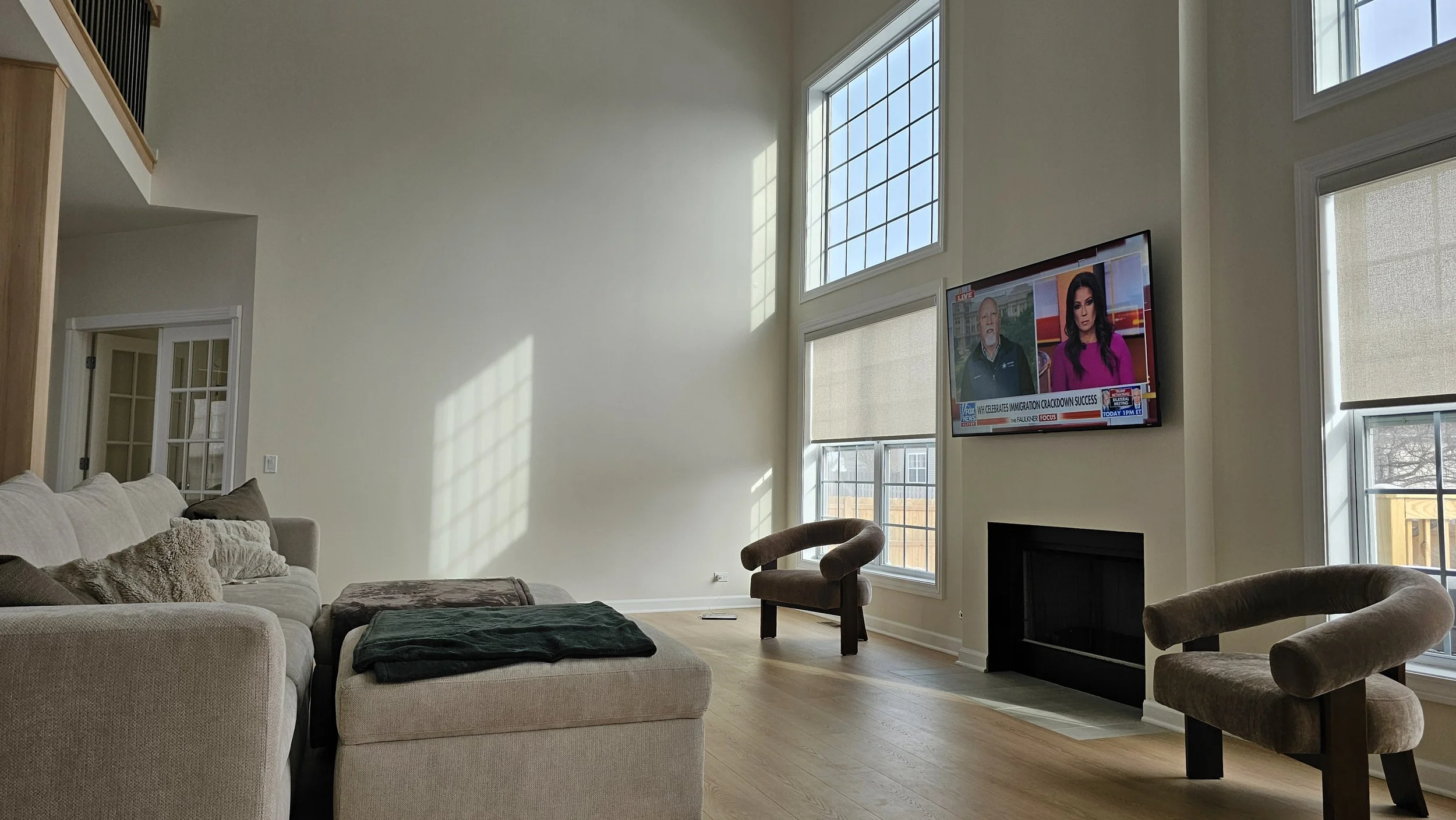 A spacious living room with a light-colored sectional sofa, two brown armchairs, a flat-screen TV on the wall showing a news broadcast, large windows with blinds, and hardwood floors.
