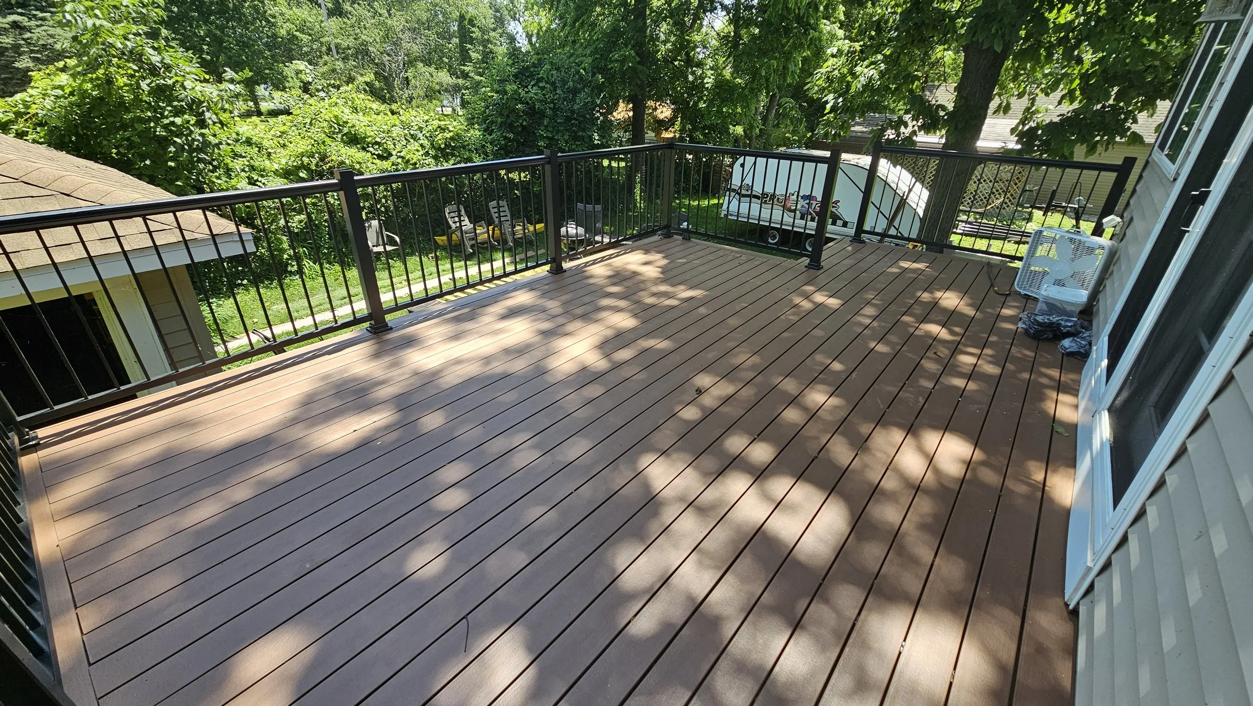 Empty outdoor deck with wood planks, black metal railing, and shadows of trees, adjacent to a house with sliding glass doors and outdoor furniture, in a lush backyard.
