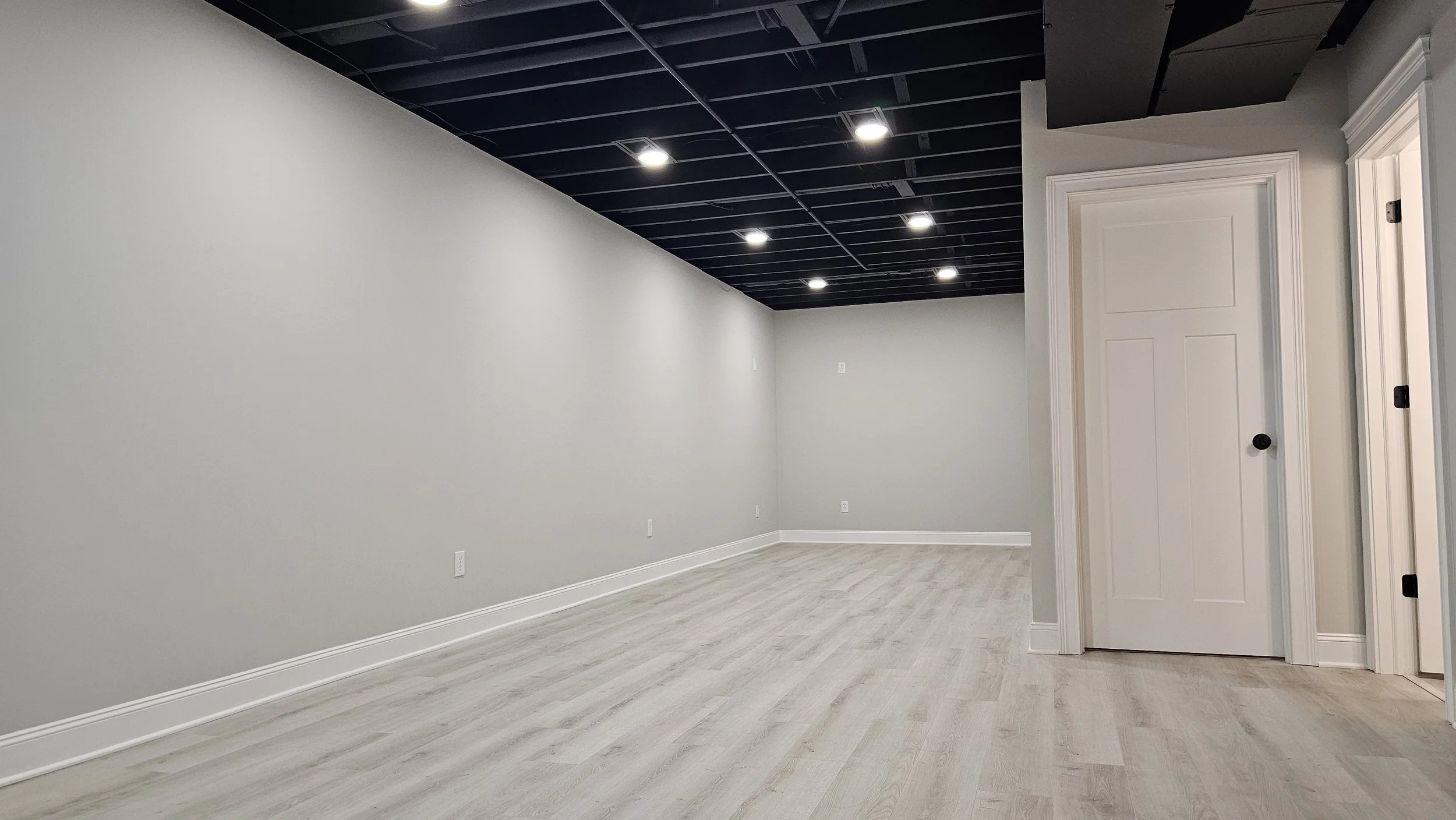 Empty room with light-colored wooden floor, plain gray walls, and recessed ceiling lights. There are two white doors with black doorknobs and crown molding details.