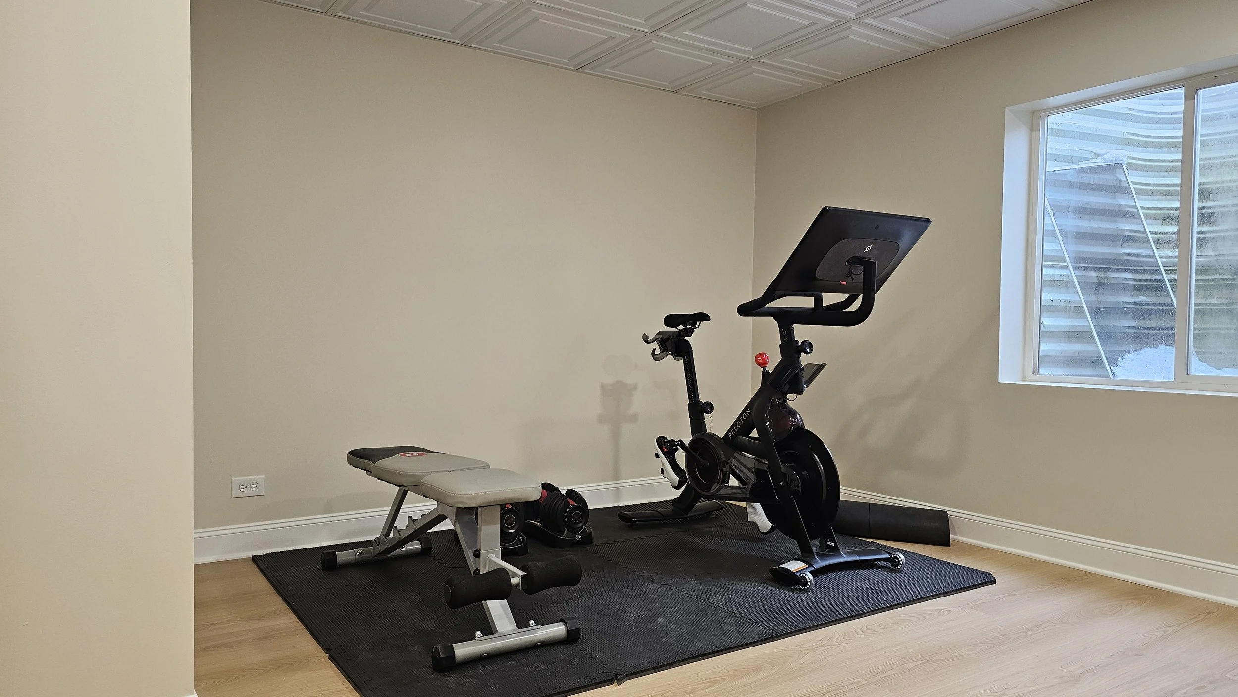 Home gym with a weight bench and an exercise bike near a window in a room with light-colored walls and wood floor.