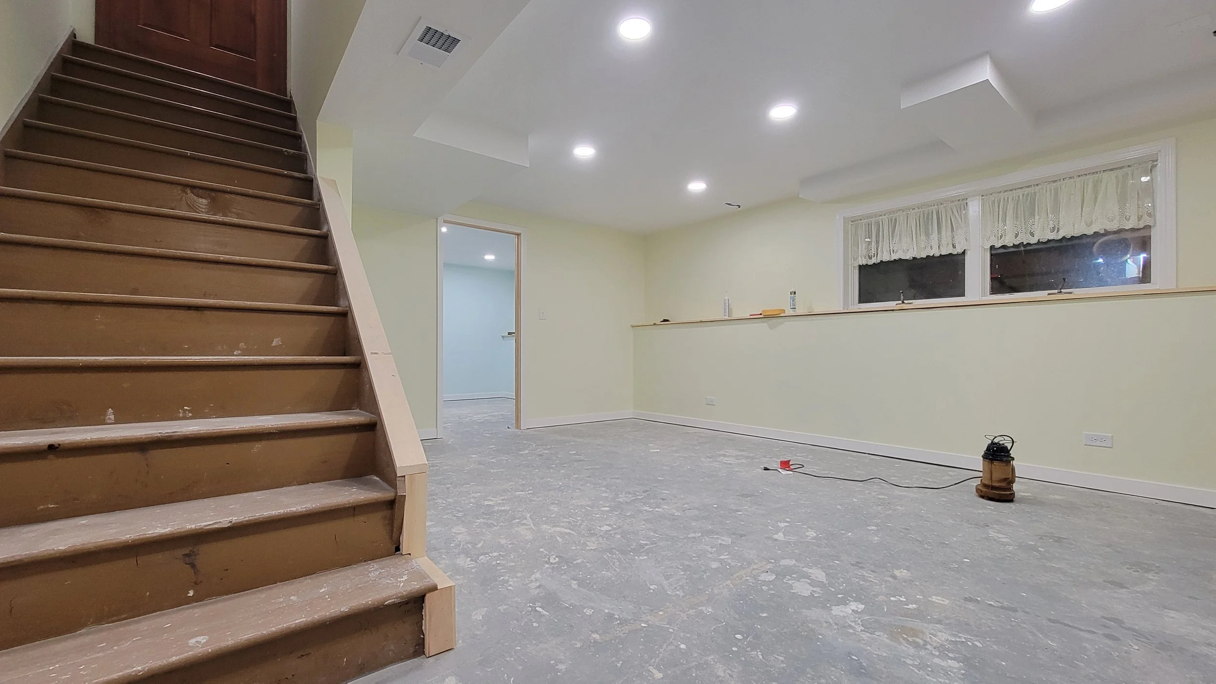 Interior of a room under renovation with a staircase on the left, a large window with curtains, and a partially finished floor.