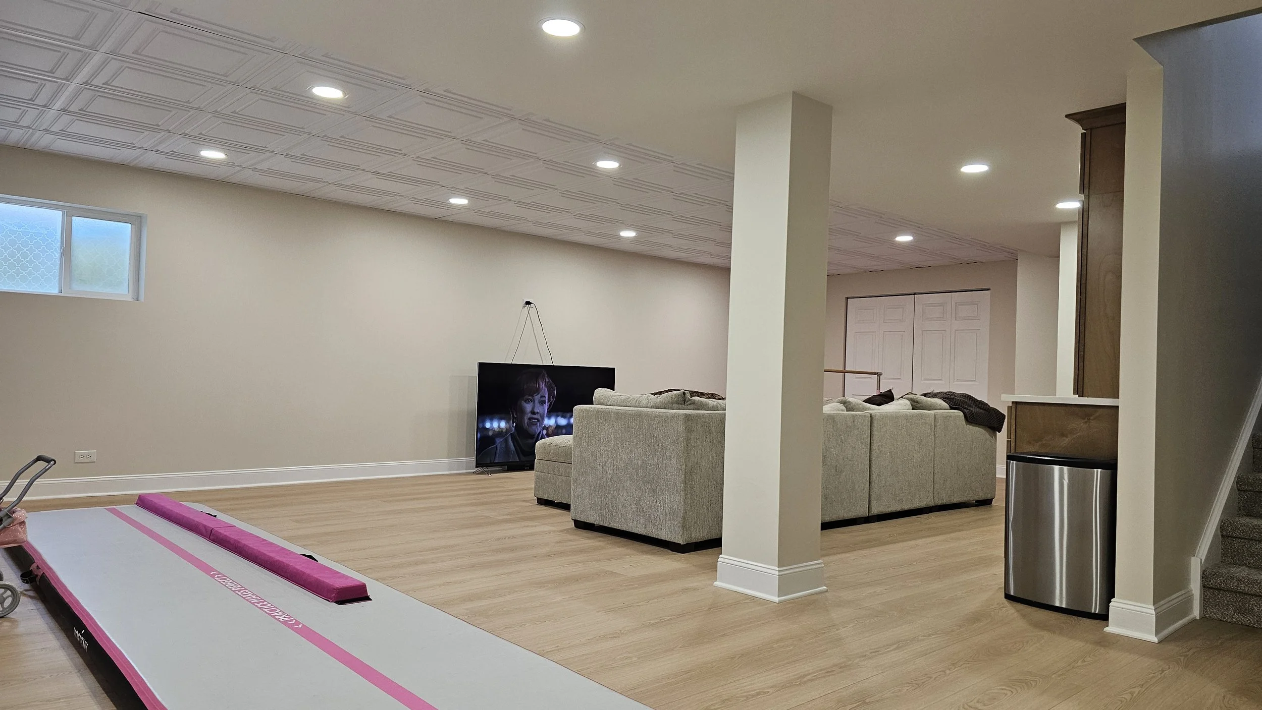 Empty basement living area with a television, light-colored sectional sofa, and a pink and white gymnastics mat on light wood flooring. The room has white walls, a small window, multiple ceiling lights, and a staircase on the right side.