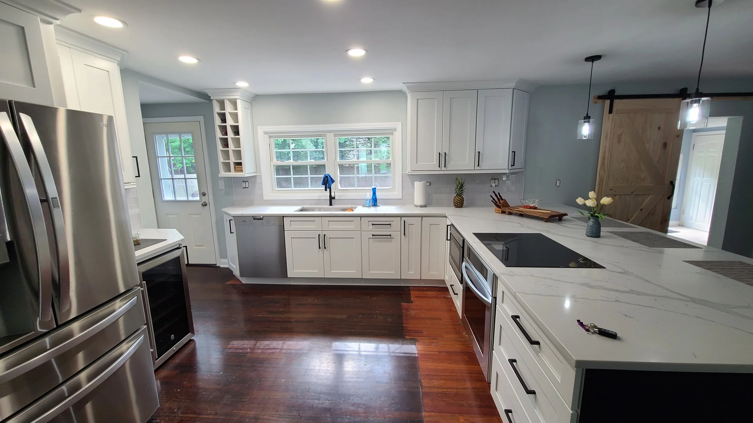 Modern kitchen with white cabinets, stainless steel appliances, a dark wood floor, and a window above the sink with outdoor greenery visible. Features include a black stovetop, a wine fridge, hanging pendant lights, and decorative plants and flowers.