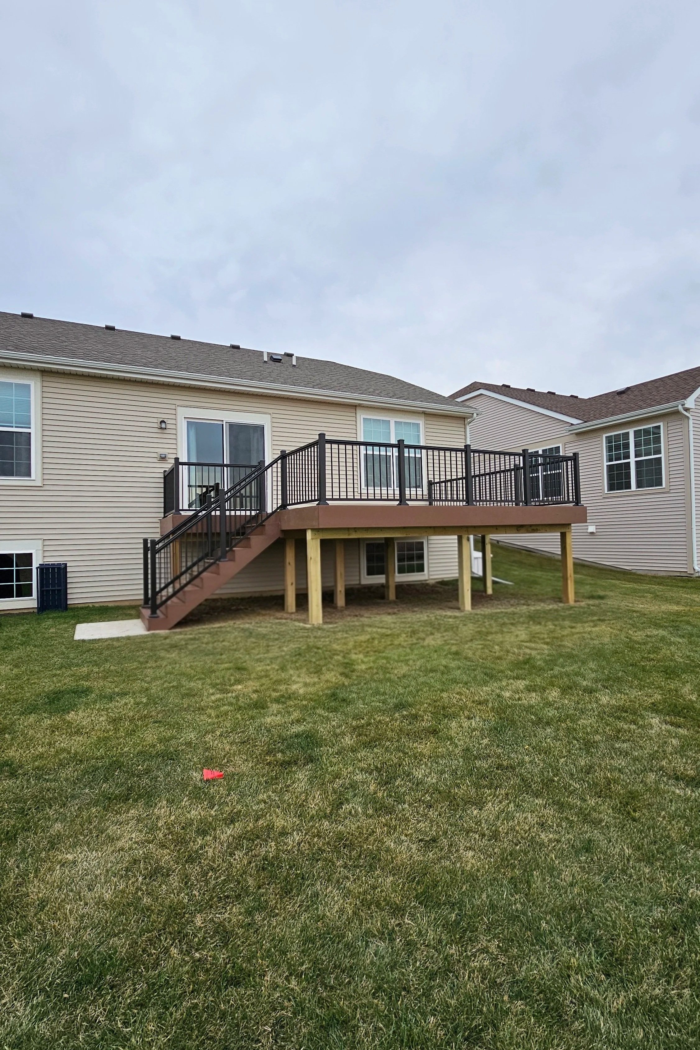 Backyard with a wooden deck attached to a beige house, accessible by stairs, with a black metal railing. The yard has green grass and neighboring houses are visible in the background under a cloudy sky.