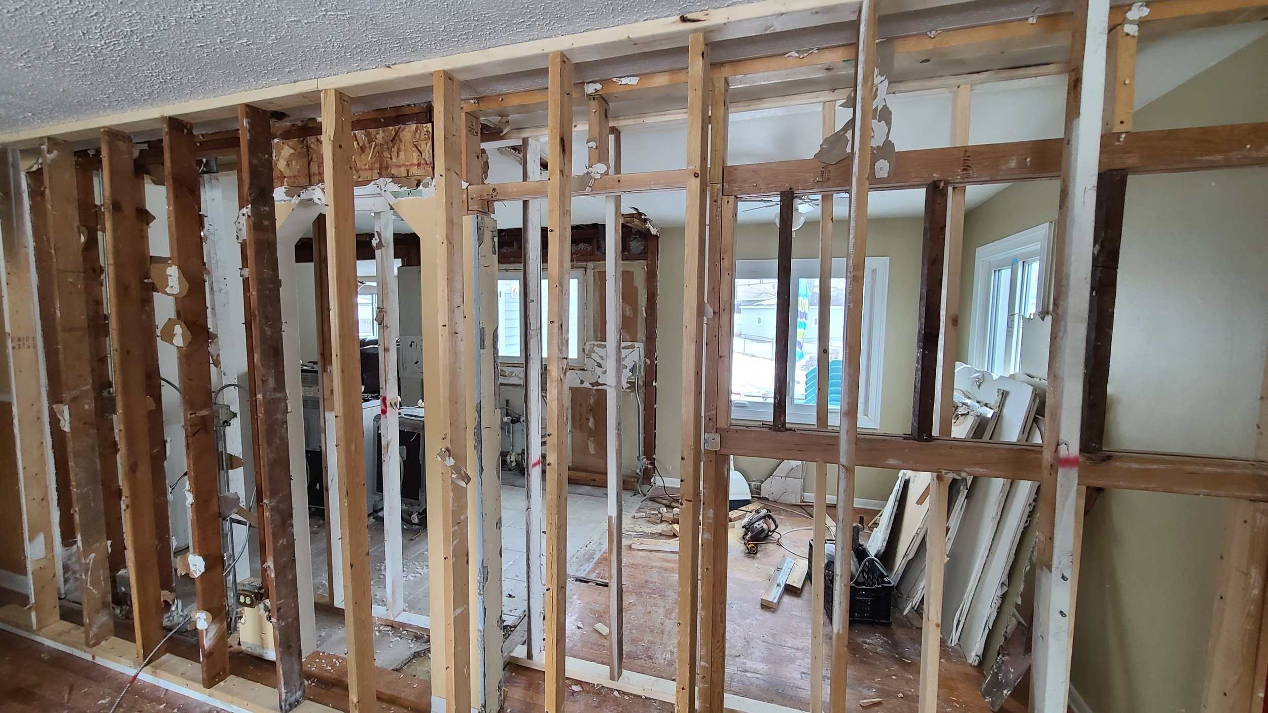 Interior of a house under renovation with exposed wooden studs and partial wall removal, showing a room with windows, construction tools, and debris on the floor.