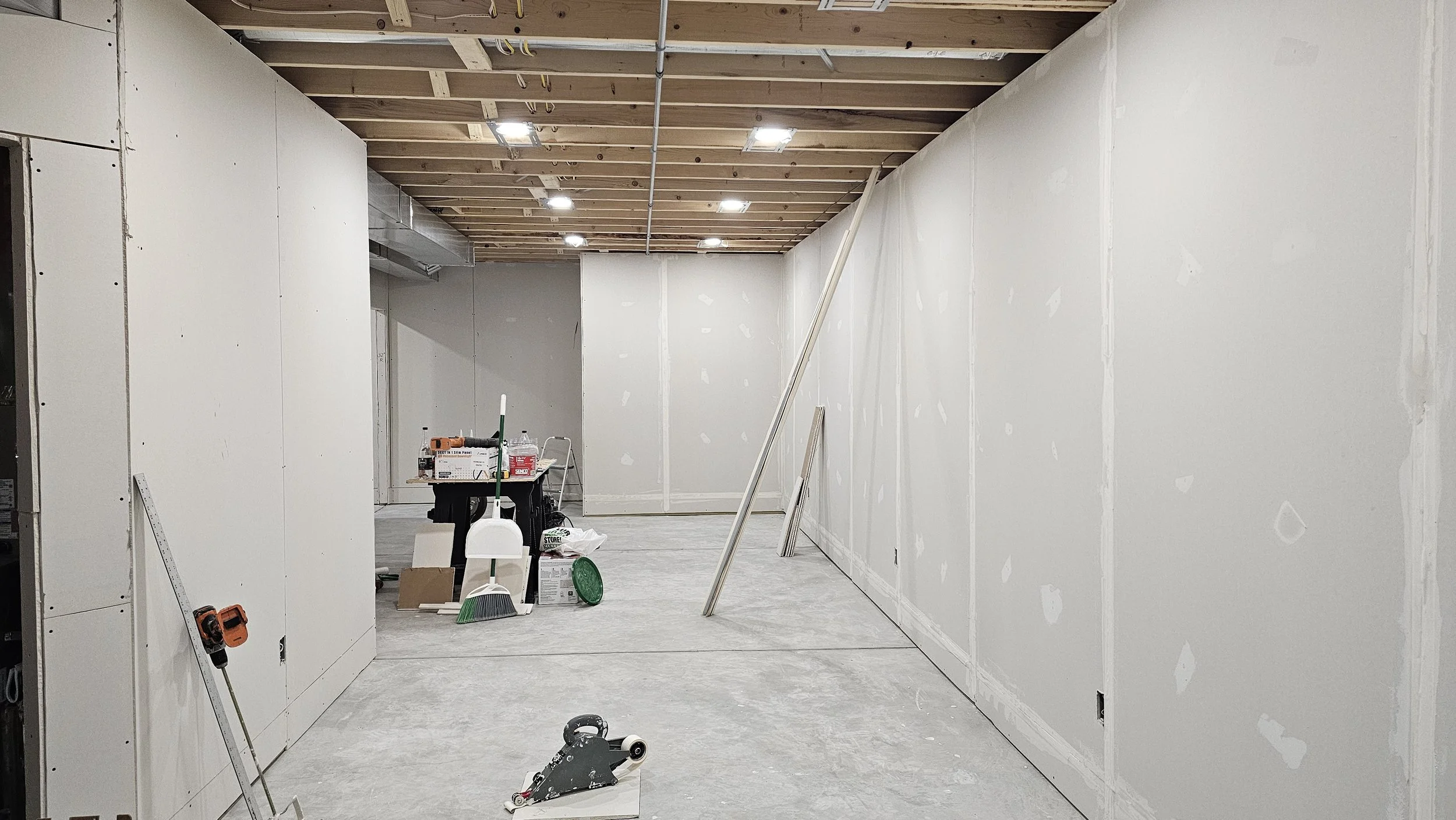 Unfinished room under construction with drywall on the walls, a wooden ceiling with exposed beams, construction tools and materials scattered on the floor, and a work table in the background.