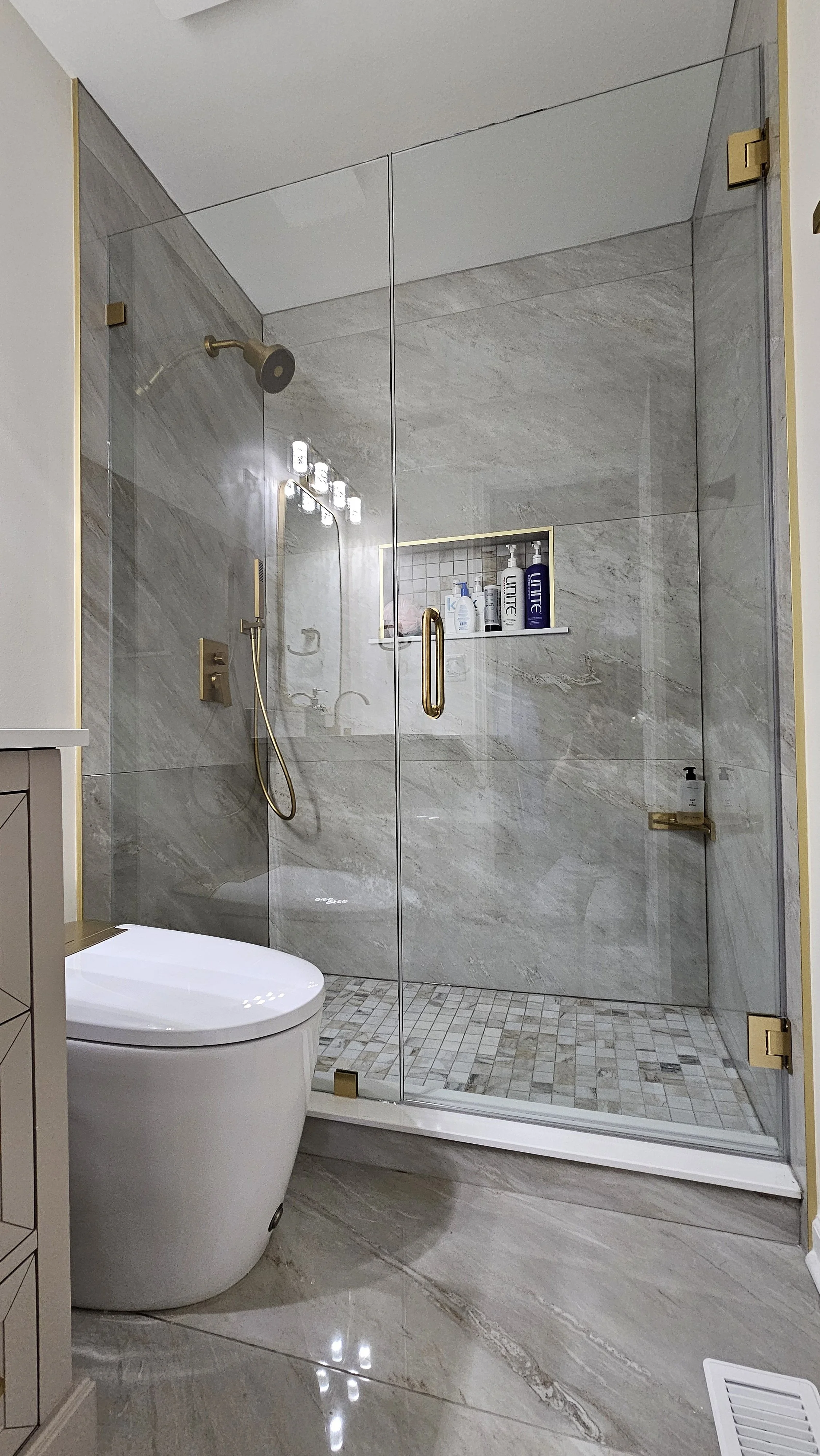 Modern bathroom with a glass-enclosed shower featuring gray marble walls and floor, gold fixtures, and a built-in niche with toiletries; a white toilet is nearby.