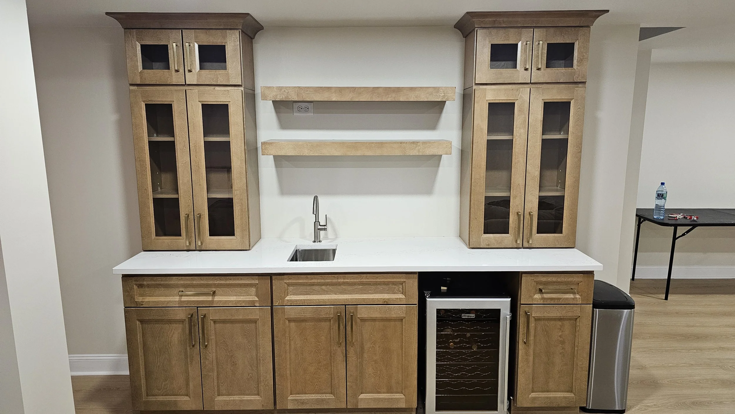 Wooden kitchen cabinets with glass doors, a white countertop with a small sink and faucet, two floating shelves, a wine cooler, and a trash bin in a room with light wood flooring.