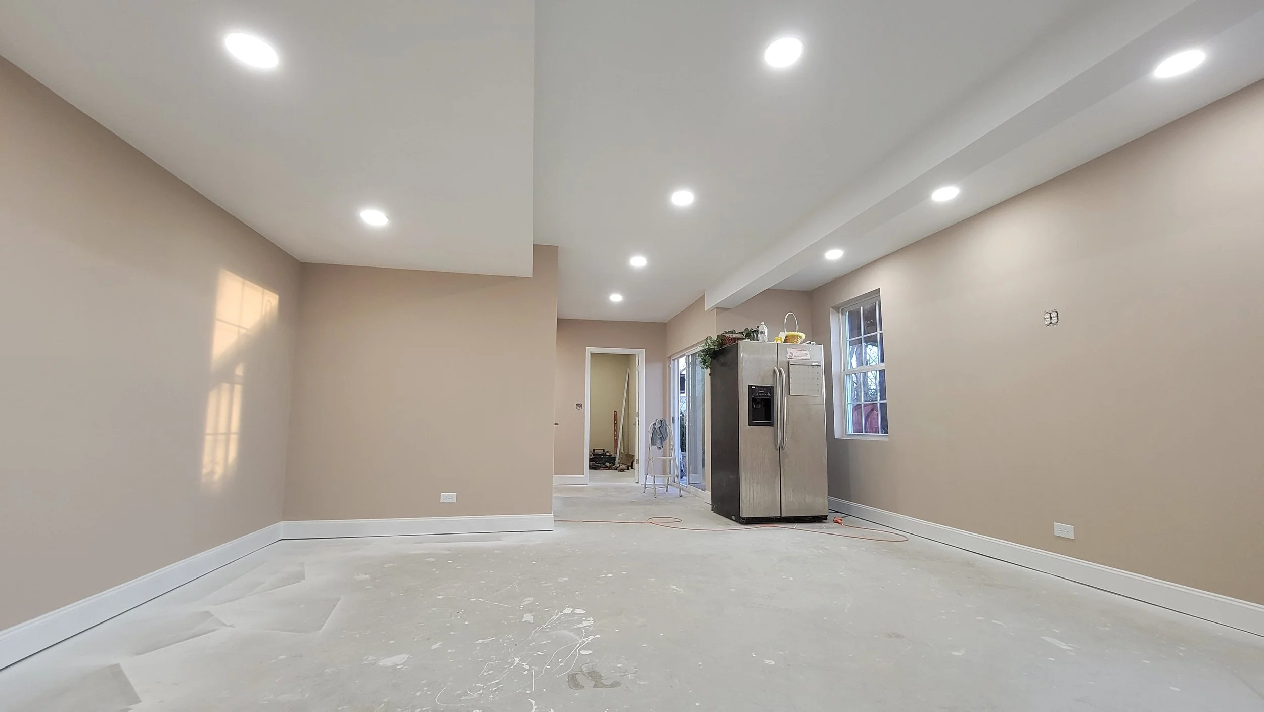 Empty living room with beige walls, white baseboards, and recessed ceiling lights, under construction with a gray refrigerator, windows, and construction tools and materials