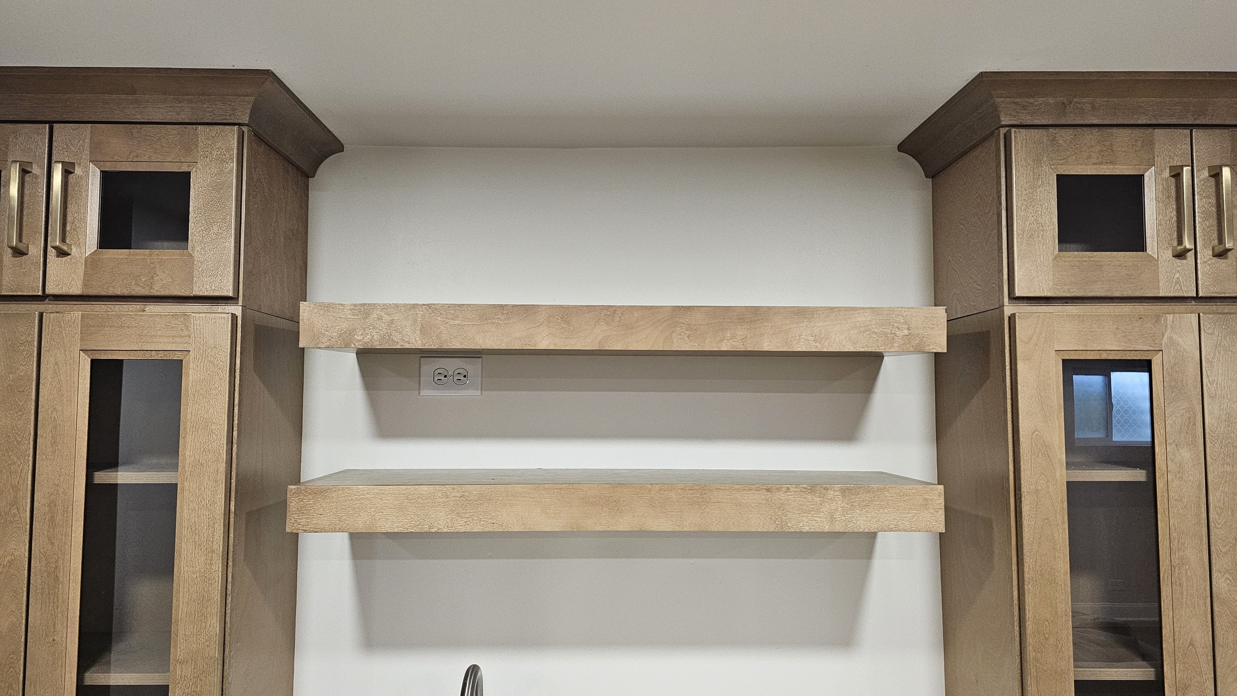 Empty kitchen wall with two wooden shelves and two tall wooden cabinets with glass doors.