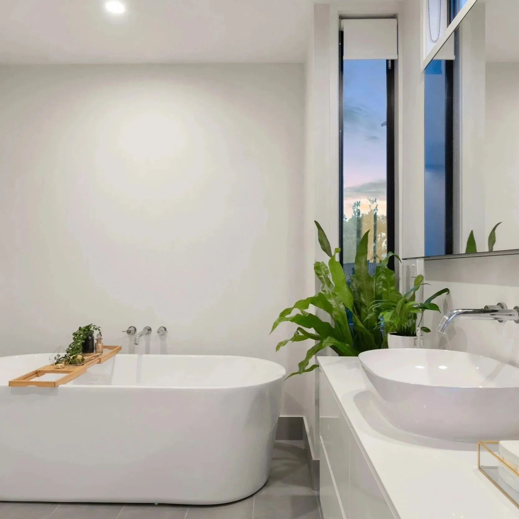 Modern bathroom with a white freestanding bathtub, a wooden bath tray with plants, and a white vanity with a vessel sink and a large potted plant, with tall windows showing a sunset sky.