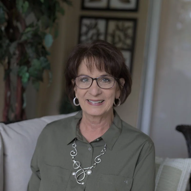 A woman with short brown hair and glasses, wearing a green shirt and a silver necklace, smiling and sitting on a light-colored couch in a well-lit room.
