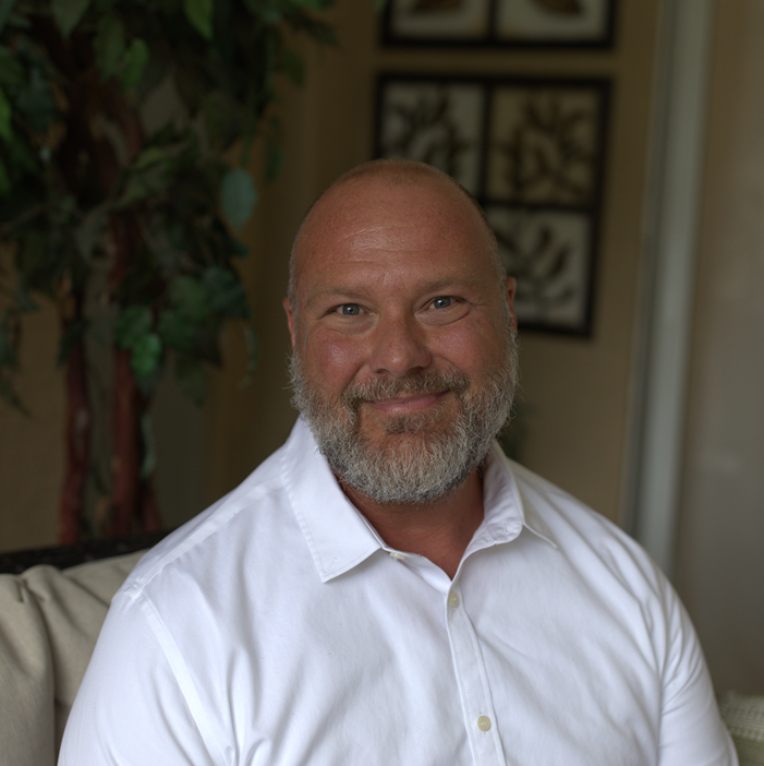 A middle-aged man with a bald head and gray beard, wearing a white button-up shirt, smiling and sitting indoors with framed artwork and a plant in the background.
