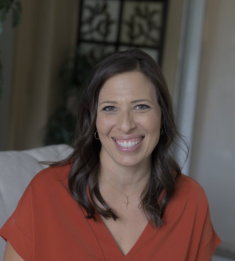 A woman with shoulder-length brown hair and blue eyes, smiling and wearing a rust-colored top.