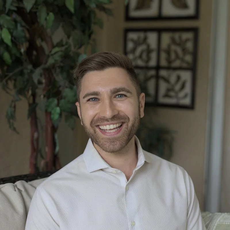 Headshot of a smiling man with brown hair and beard, wearing a white collared shirt, sitting indoors with houseplants and framed artwork in the background.