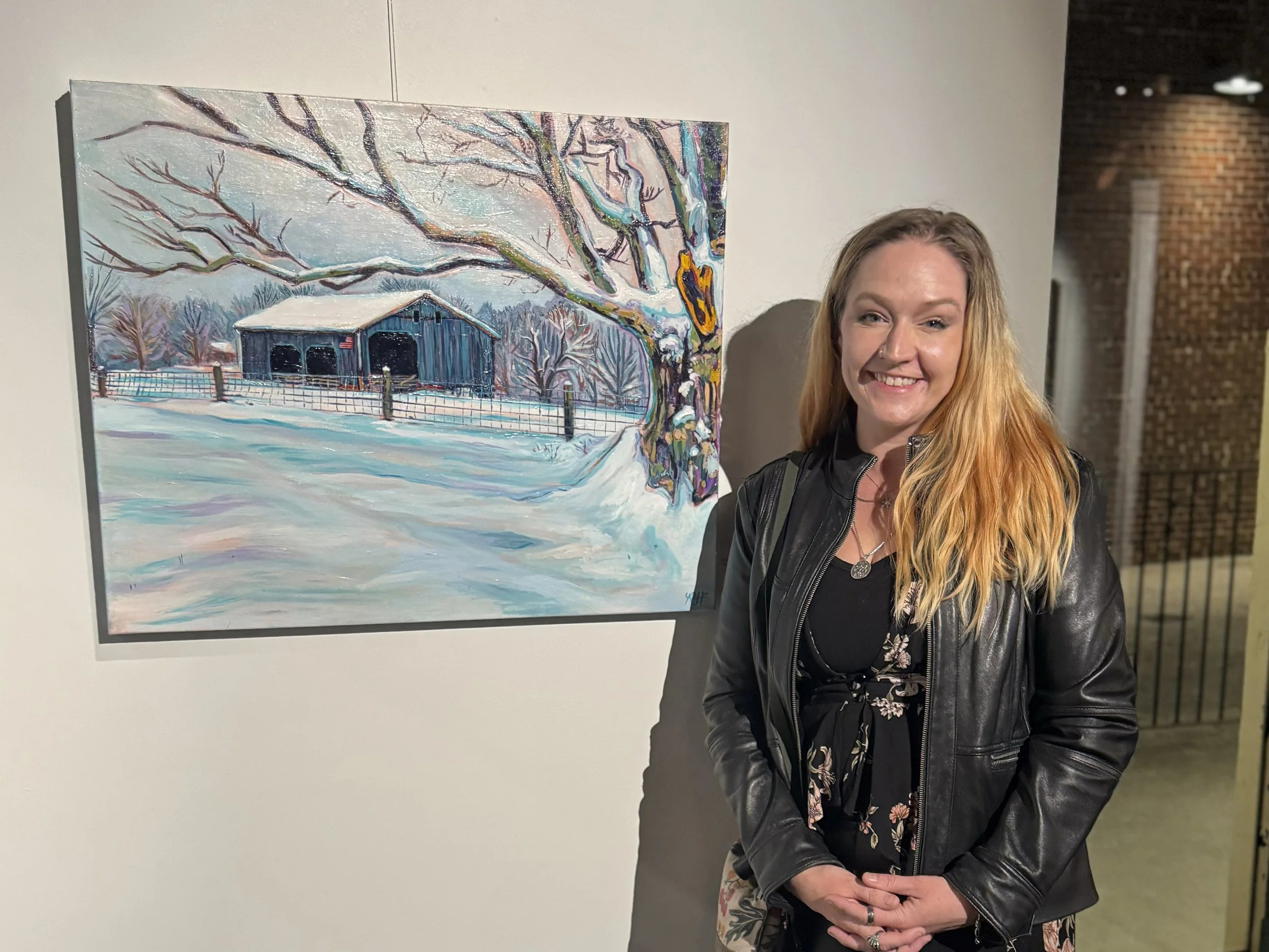 Rachel J Haungs a white woman with blonde hair in a leather jacket standing in front of her 30 inch wide 24 inch tall acrylic painting in vibrant hues of a tree with a wound from a limb cut off in the foreground, cabin in the center in a snow scene.