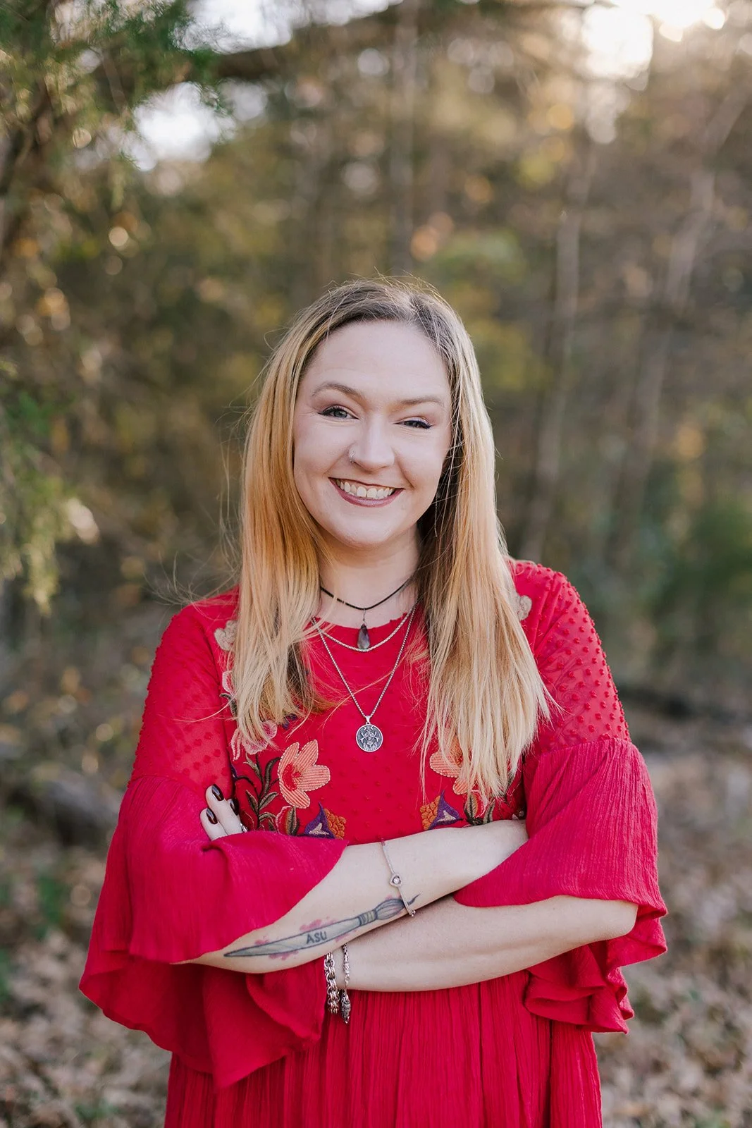 Photograph of Rachel J Haungs, a white female in her mid thirties with blonde hair and a red dress with bell sleeves on and her arms crossed standing in front of the forest.
